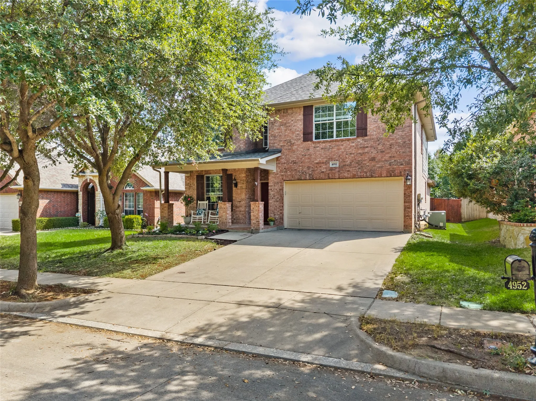 Brick exterior with a covered porch, an attached garage, a concrete driveway, and mature landscaping
