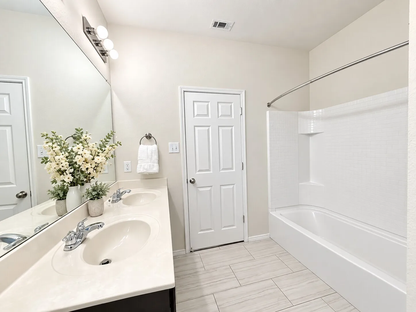 Dual vanity with chrome fixtures and integrated sinks, a full-width mirror, a three-light vanity bar fixture, a white shower-over-tub combination with a tiled surround, and light-toned tile flooring