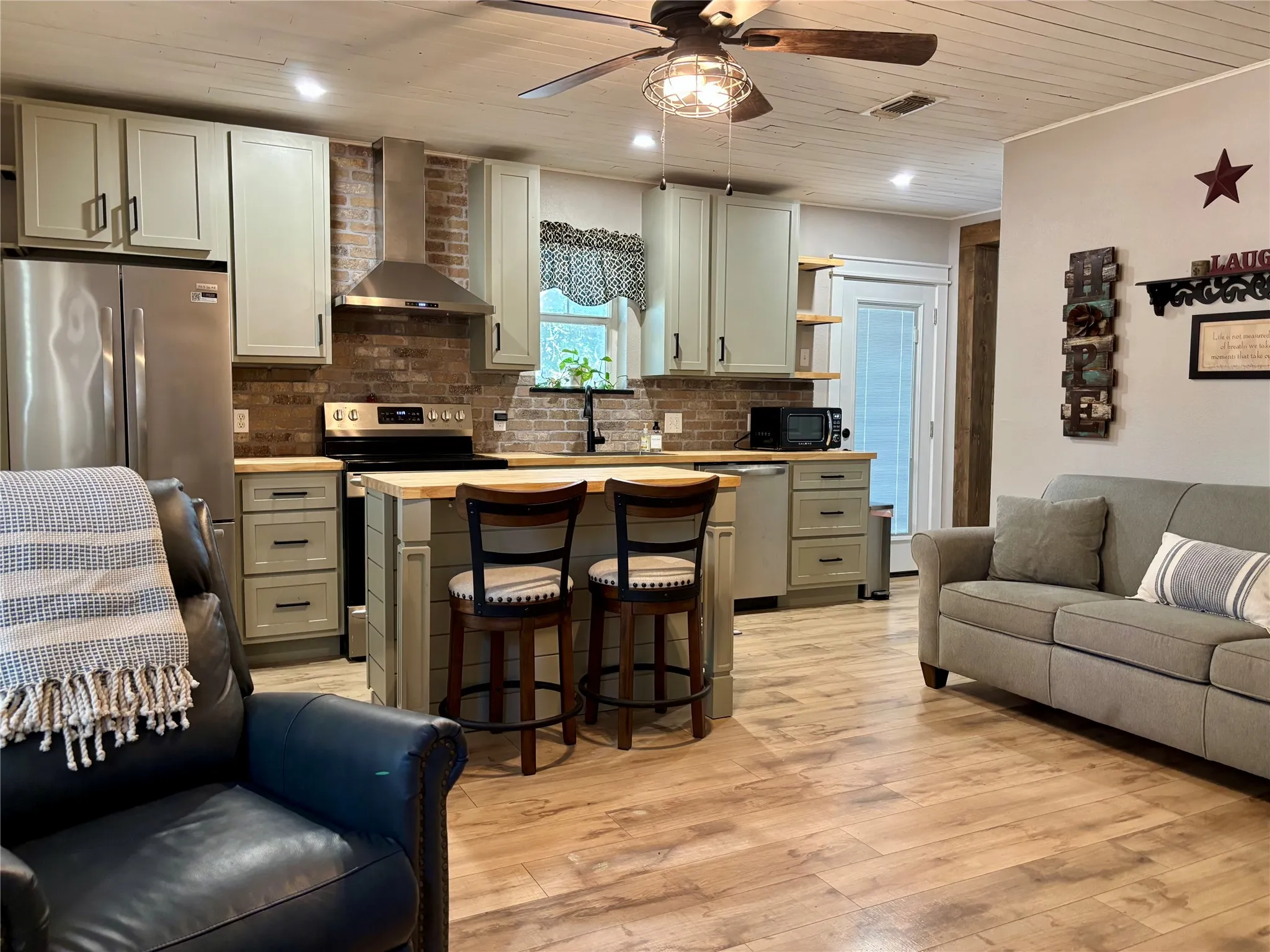 Kitchen featuring wood-finish flooring, brick backsplash, light-toned cabinetry, ceiling fan, shiplap ceiling. The door leads to the side porch with fenced yard.