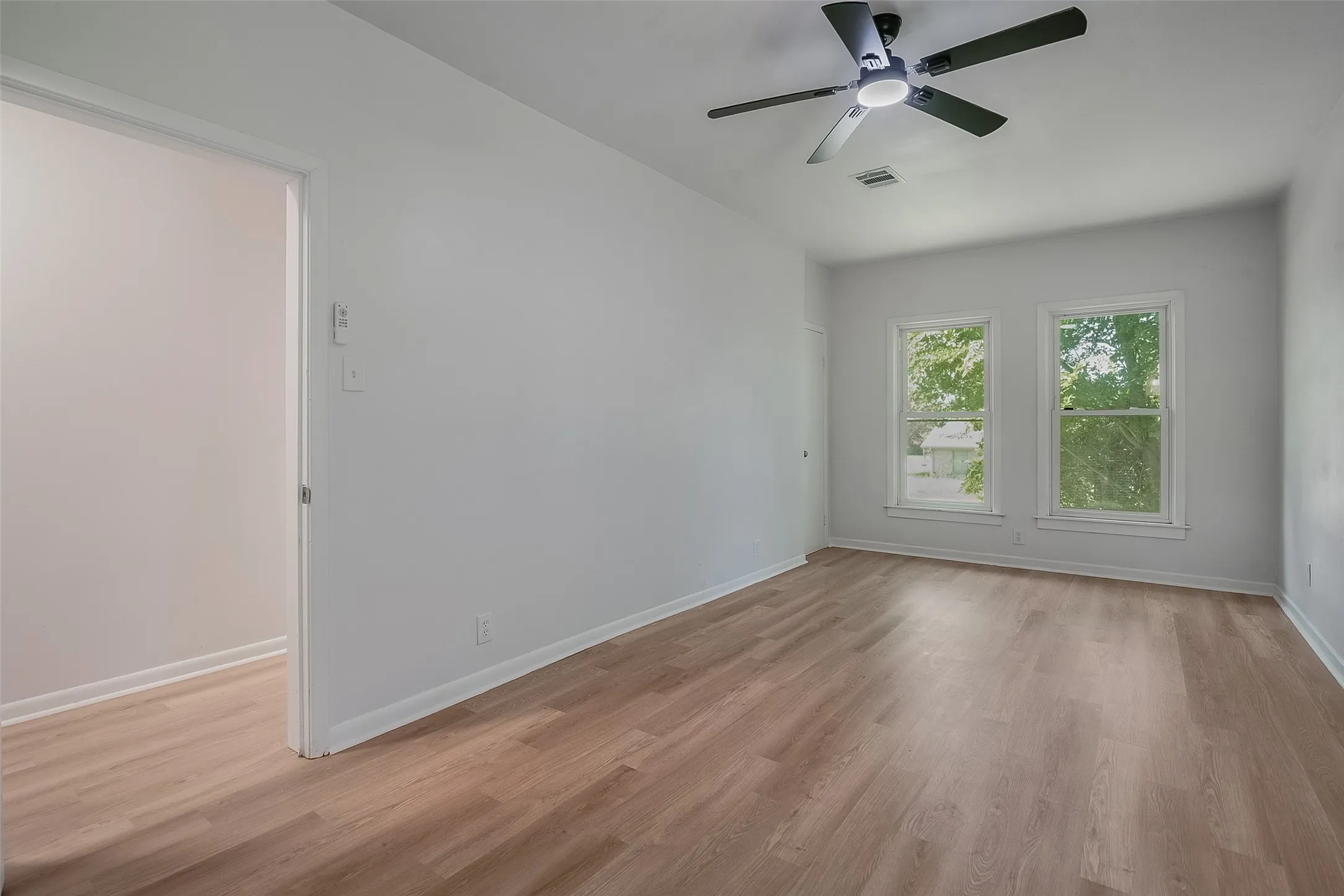 Wood-finish flooring, white painted walls, two windows with white trim, and a ceiling fan with integrated lighting