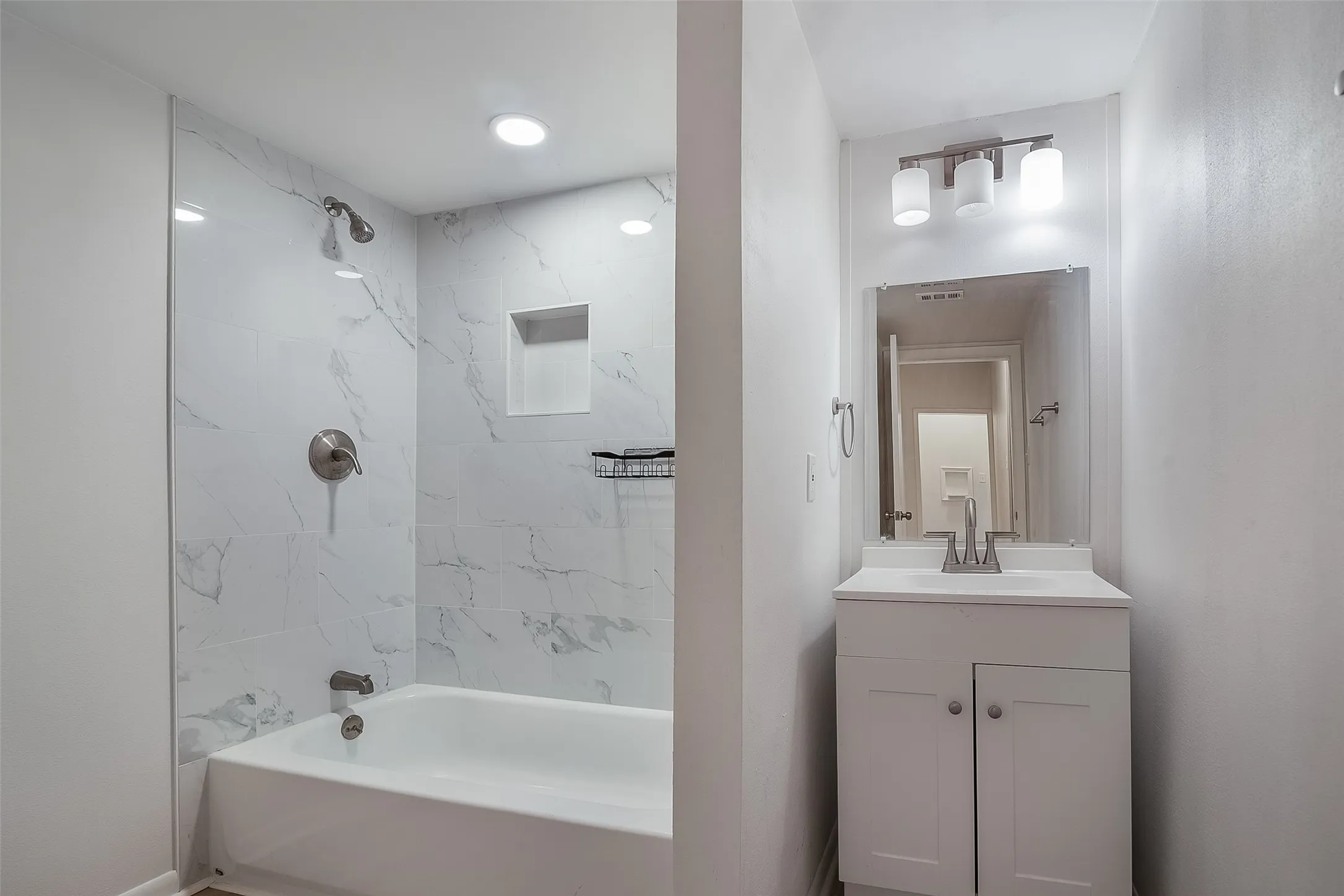 White vanity with chrome faucet and lighting, framed mirror, and a shower-tub combination featuring white tile with gray veining and a recessed niche