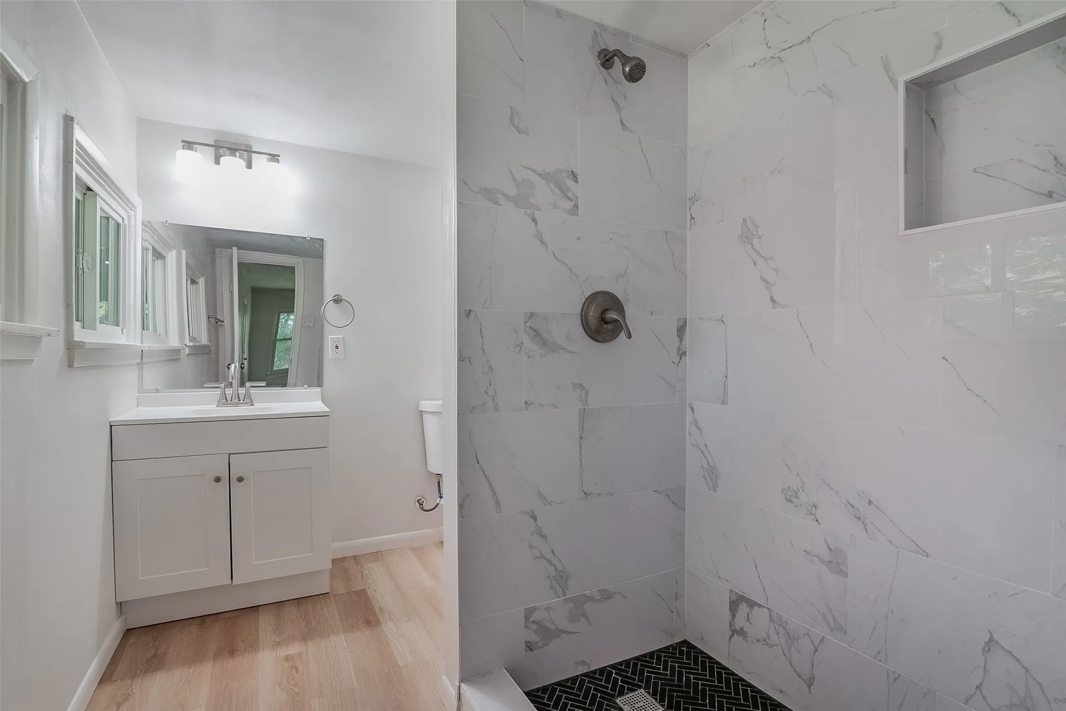White vanity with a single sink, light wood-finish flooring, and a shower with white tile walls and a dark herringbone tile floor