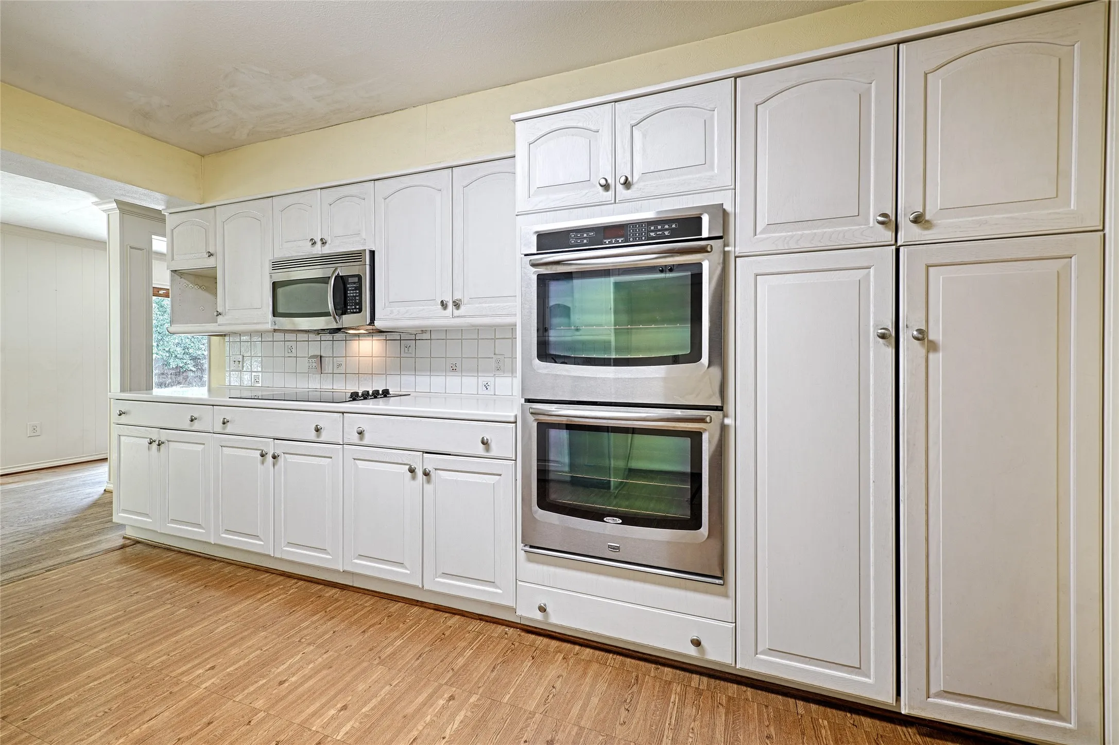 White cabinetry, wood-finish flooring, stainless steel double ovens, stainless steel microwave, and a white tile backsplash
