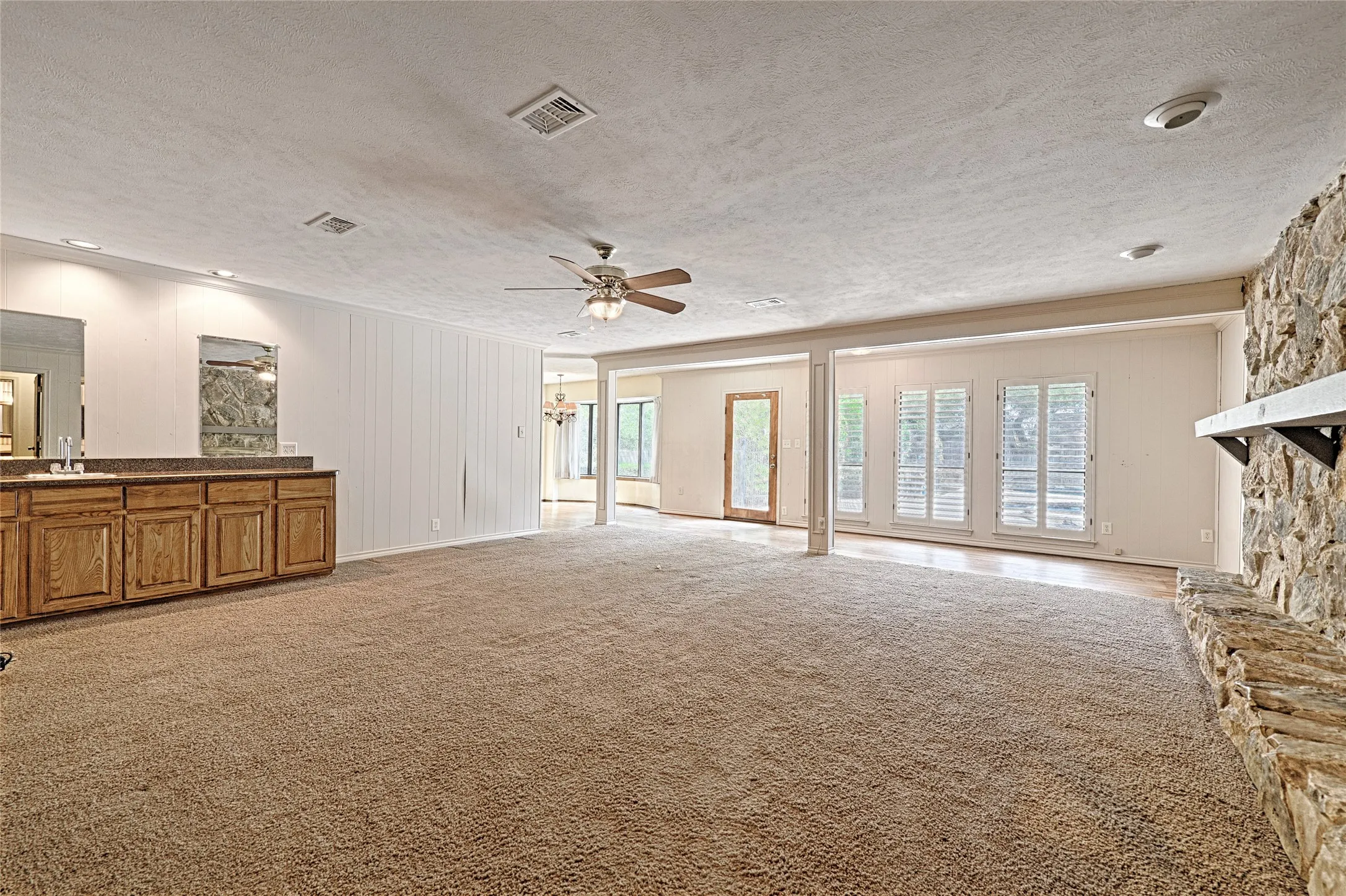 Spacious room featuring carpet flooring, a stone-clad fireplace with a mantel, built-in cabinetry with a countertop and mirror, multiple windows with plantation shutters, and recessed lighting