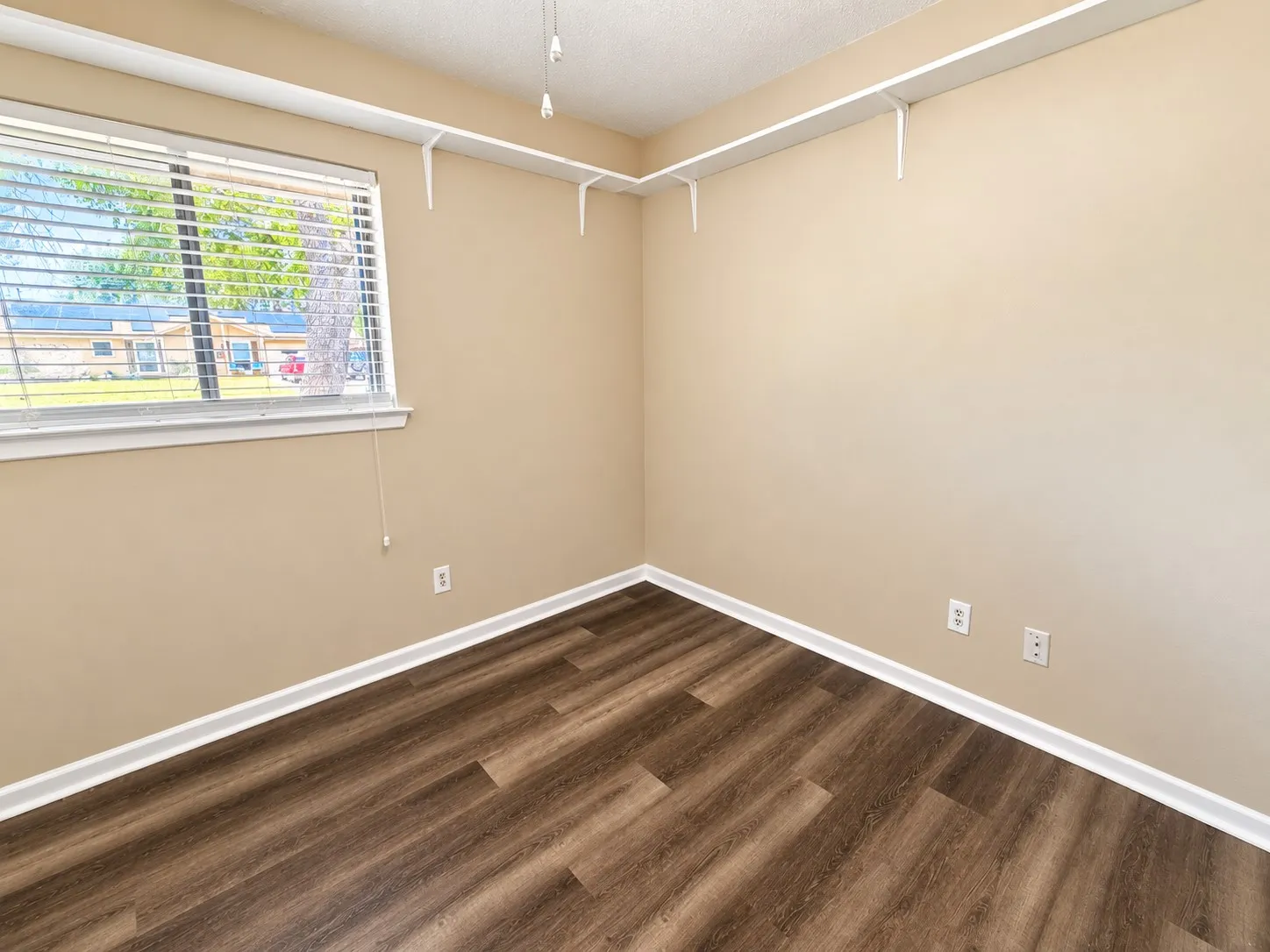 Wood-finish flooring with white trim, a window featuring horizontal blinds, and corner-mounted shelving
