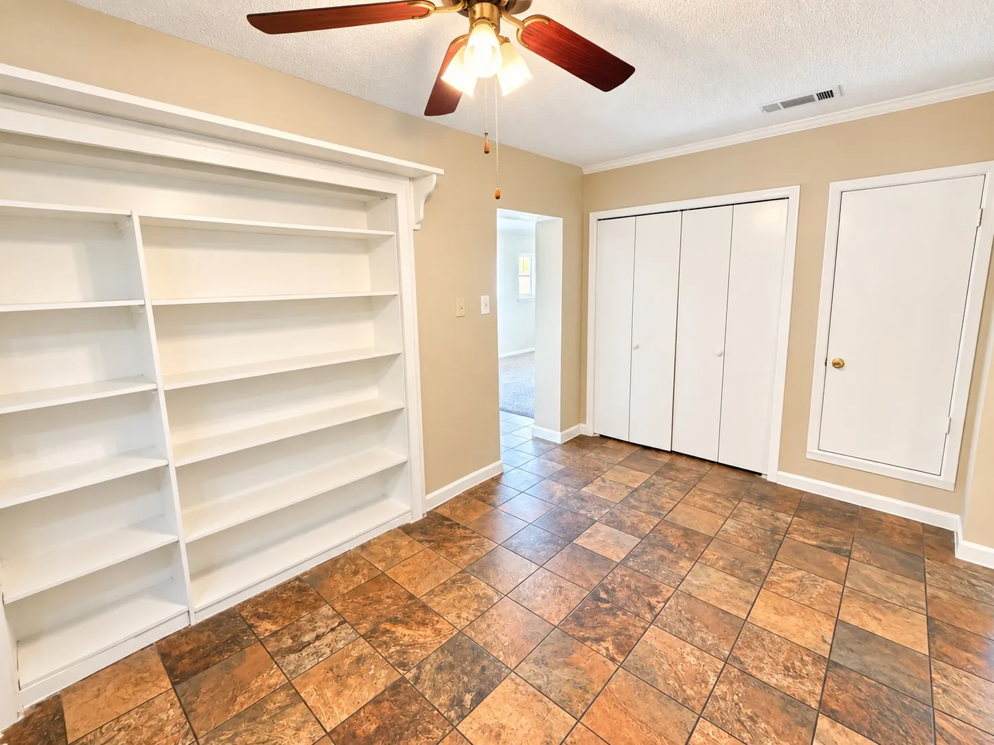 Built-in white shelving, wood-finish ceiling fan, bi-fold closet doors, and multi-tone square floor tiles