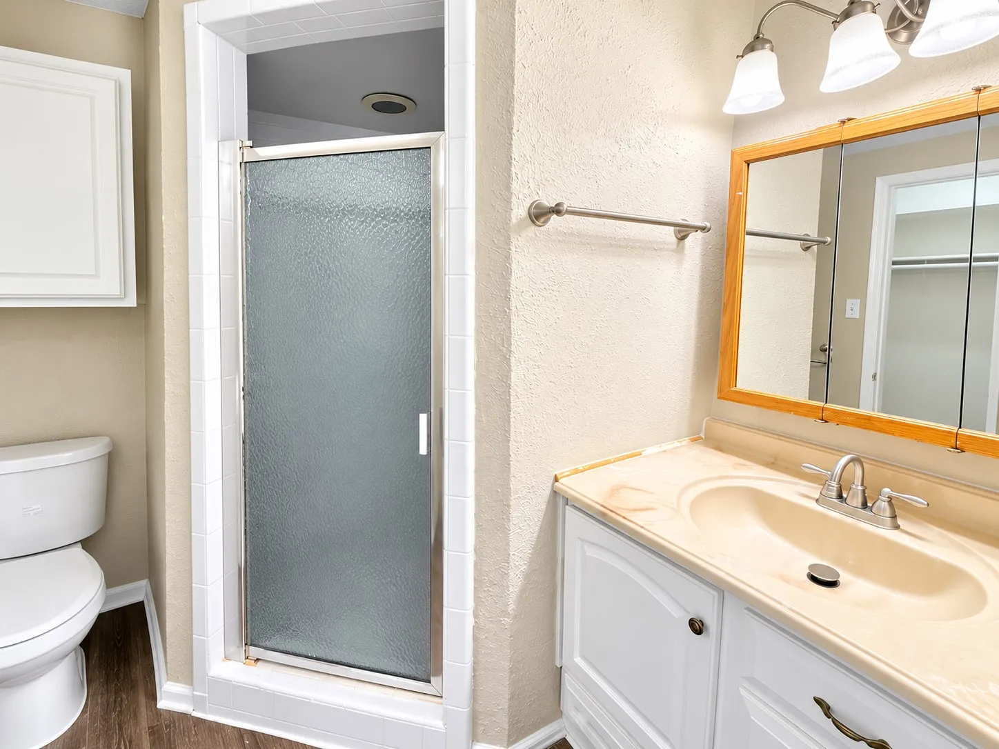Wood-finish flooring with white baseboards, shower enclosure with frosted glass door, white toilet, vanity with integrated sink and countertop, and a medicine cabinet with a wood-finish frame