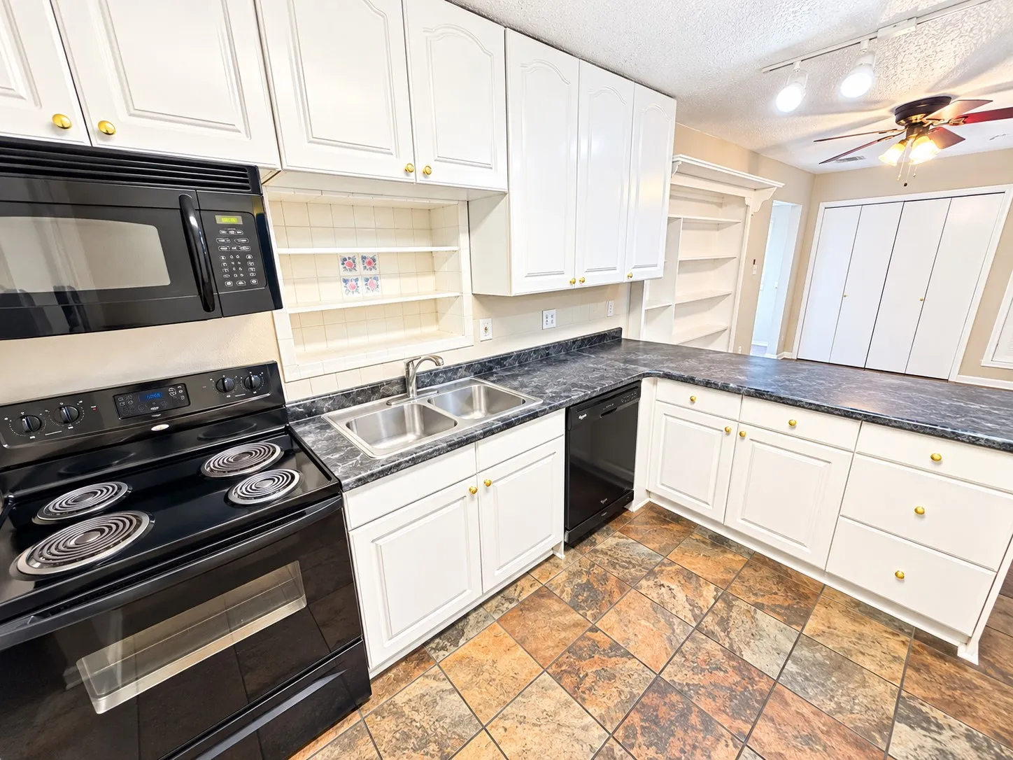 White cabinetry with gold-tone hardware, black appliances, double basin stainless steel sink, dark patterned countertops, and multi-toned tile flooring