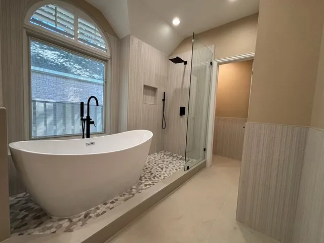 Freestanding soaking tub with matte black fixtures, an arched window featuring plantation shutters, and a walk-in shower with a glass enclosure and pebble floor