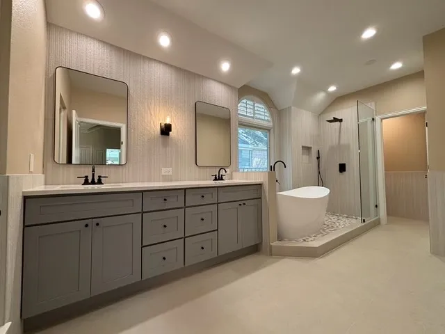 Dual vanities with matte black fixtures, free-standing soaking tub with pebble surround, glass-enclosed shower, and arched window with plantation shutters