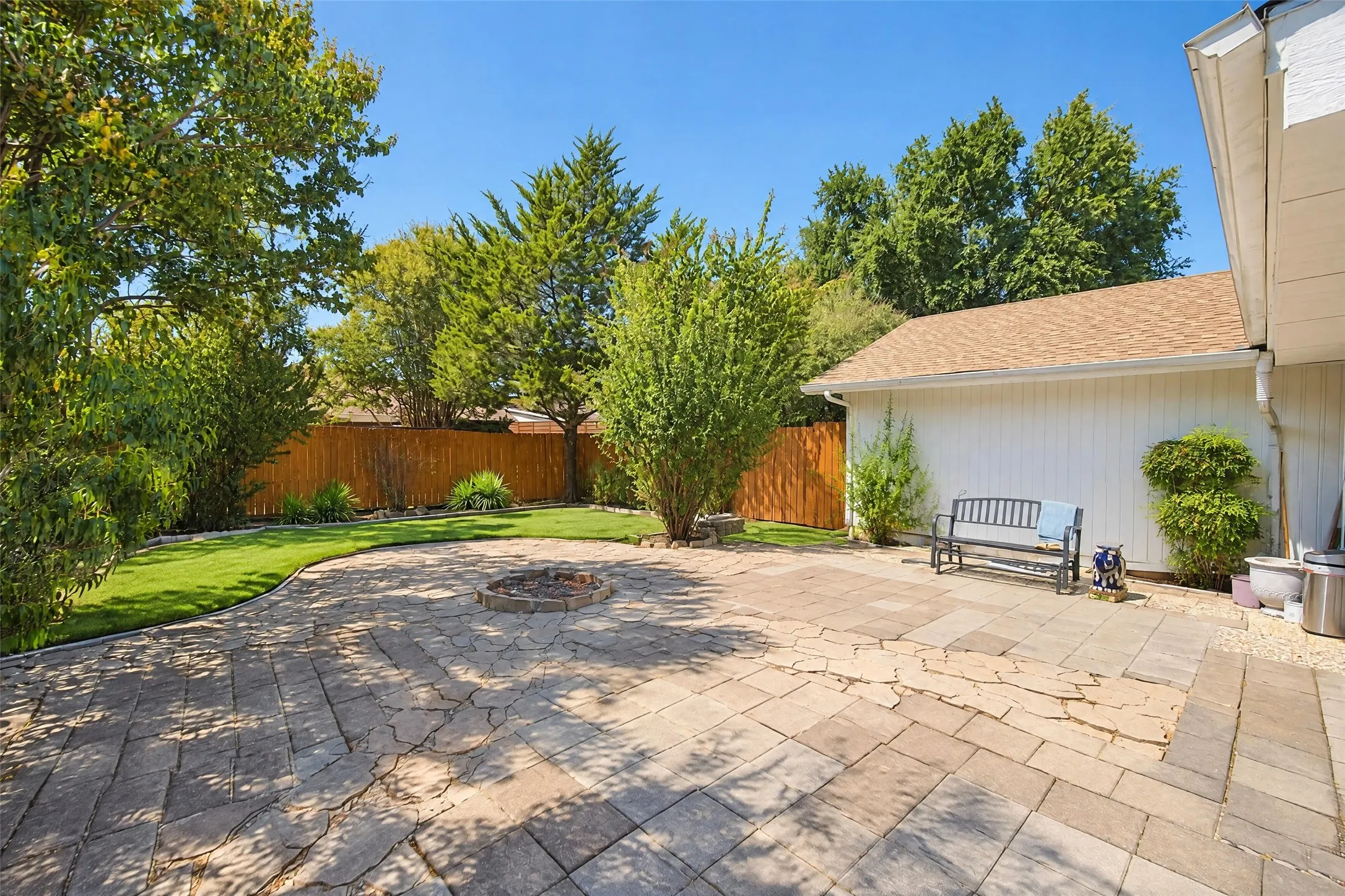 Expansive paver patio with a built-in fire pit, surrounded by lush green lawn, mature trees, and a privacy fence
