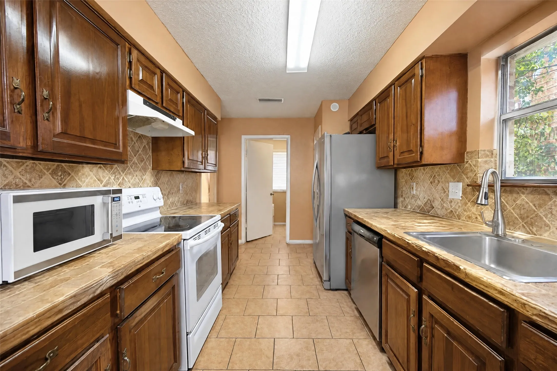 Galley kitchen featuring wood cabinetry, stone-look countertops, tiled backsplash, and stainless steel appliances