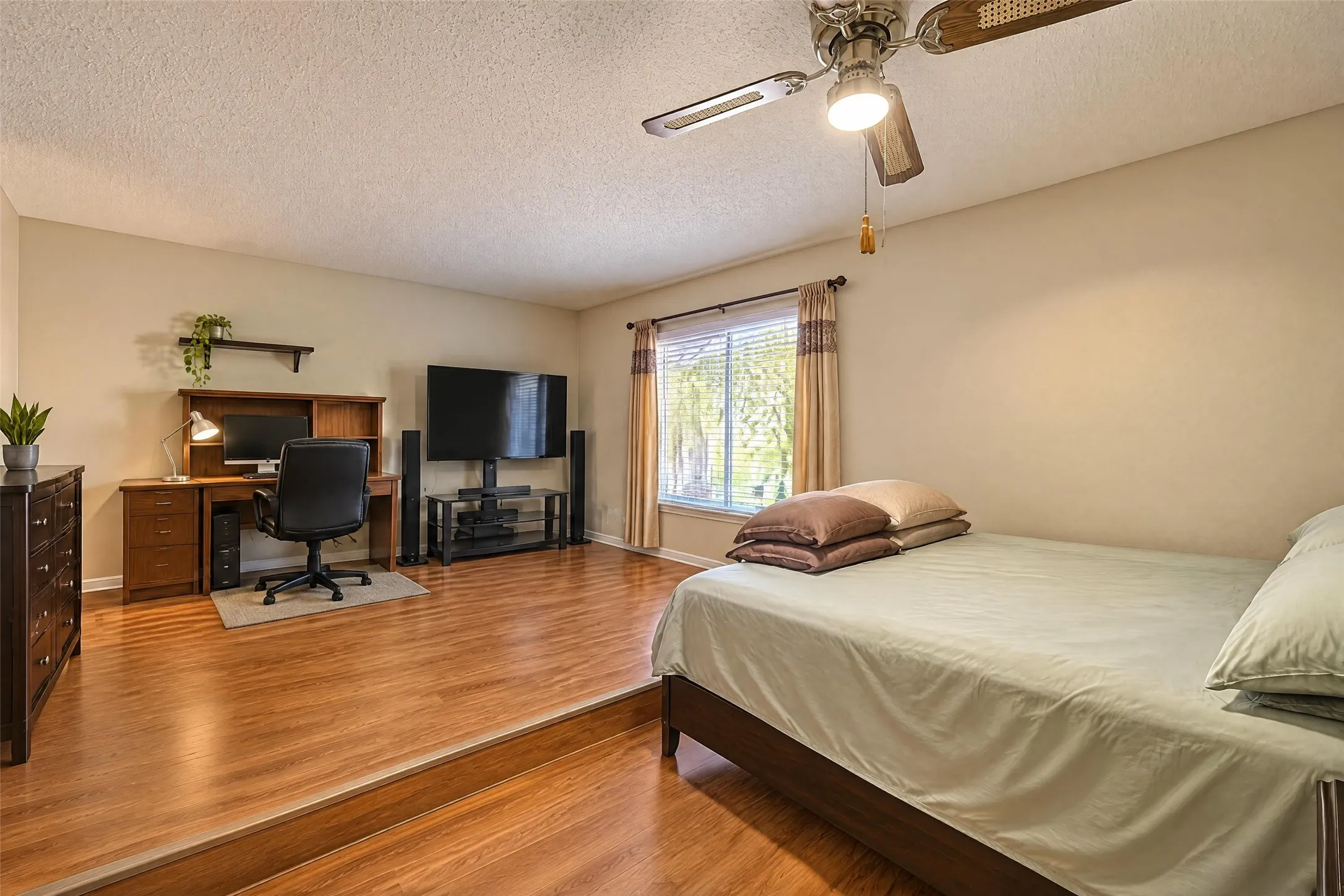 Spacious primary suite featuring hardwood flooring throughout, a queen-size bed frame, dedicated work station with desk and office chair, wood cabinetry, ceiling fan with integrated lighting, and natural light from sliding glass doors with coordinating window treatments