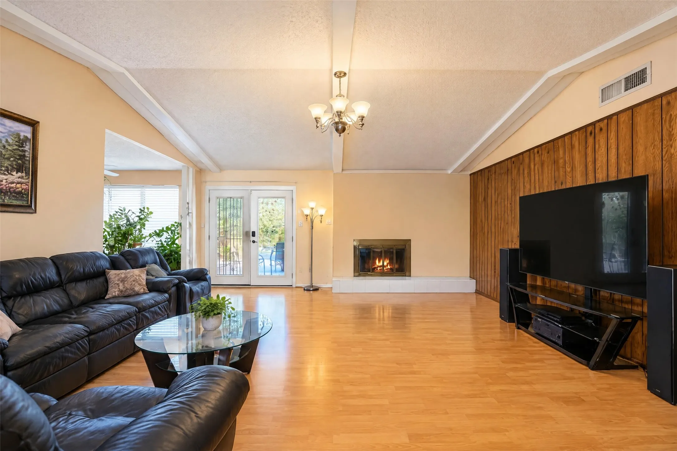 Vaulted ceiling with exposed beams, light wood-finish flooring, recessed fireplace, wood panel accent wall, and french doors