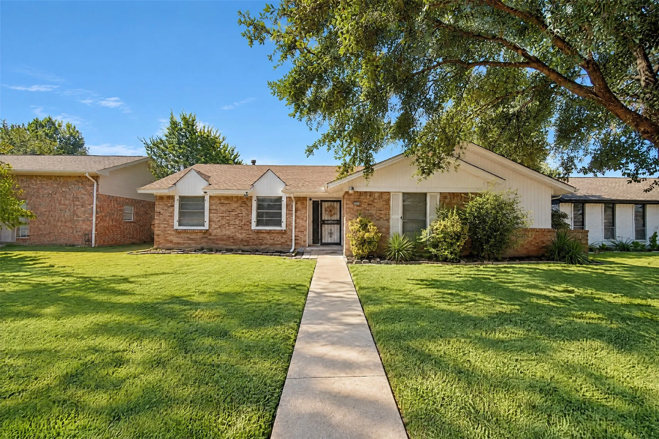 Brick exterior, gabled roofline, mature trees, concrete walkway, and landscaped front yard
