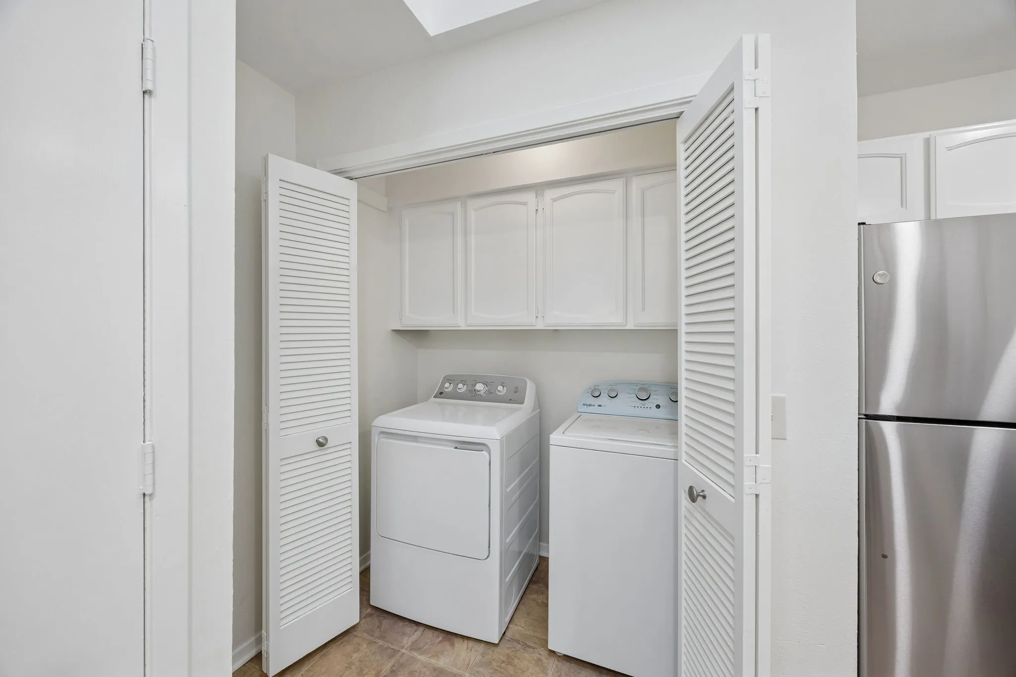 Dedicated laundry area with louvered bifold doors, upper white cabinetry, and textured tile flooring