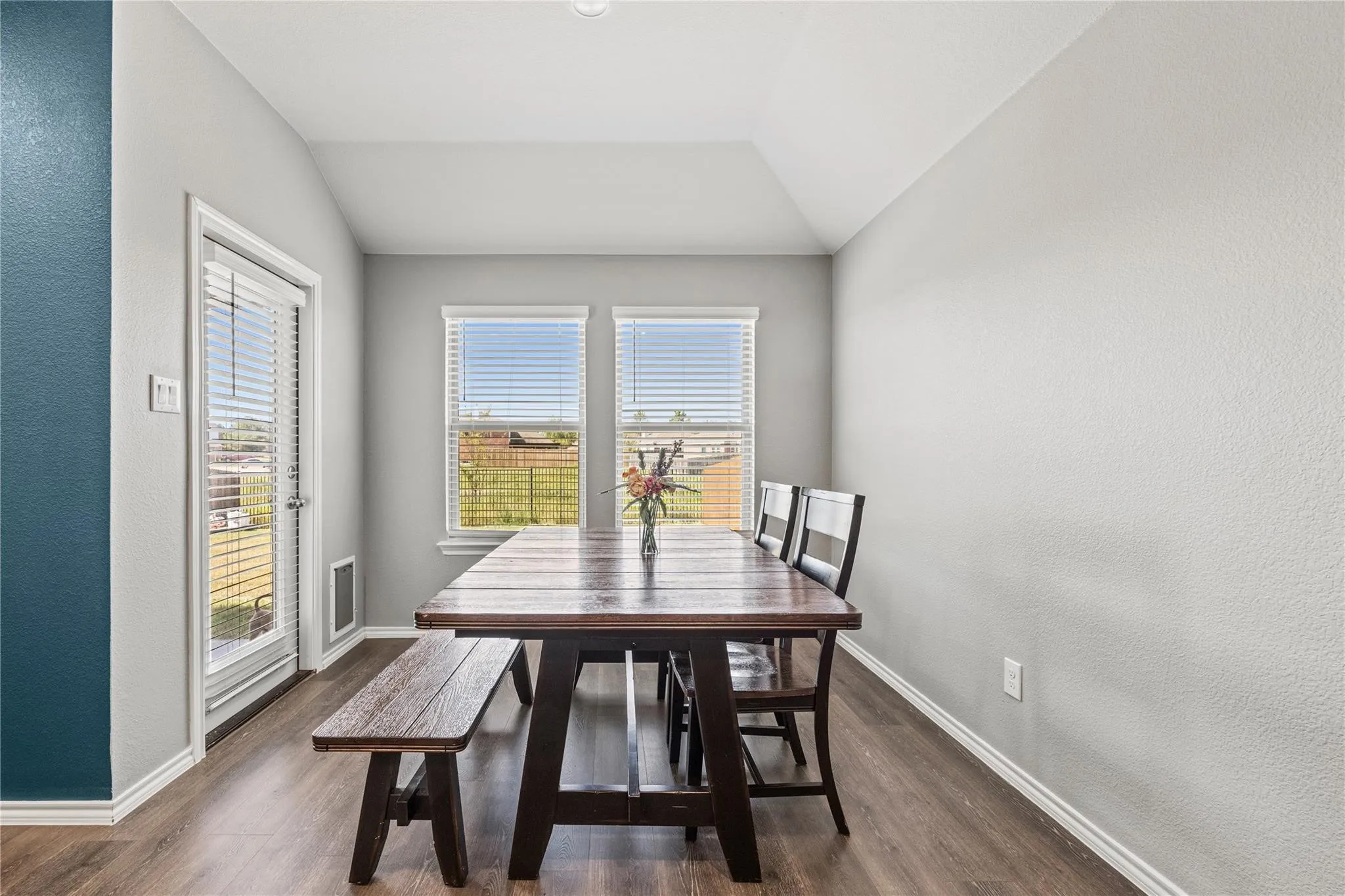 Vaulted ceiling with an egress door, dual windows featuring blinds, and wood-finish flooring