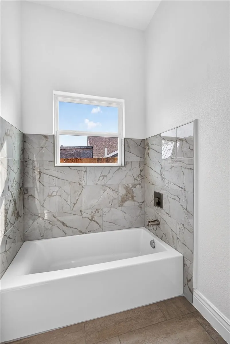 Bathtub with light-toned marble-patterned tile surround, bronze-finish fixtures, and wood-finish flooring