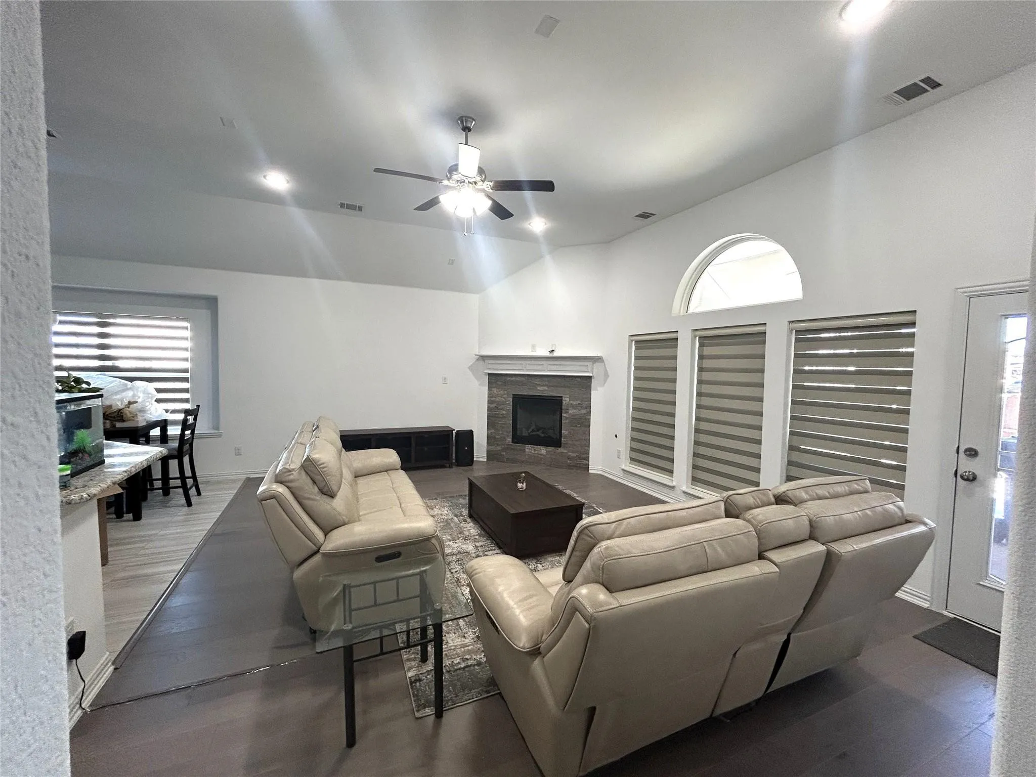 Spacious open-concept living area featuring a ceiling fan with integrated lighting, a wood-burning fireplace with surround, arched transom windows with plantation shutters, and wood-finish flooring throughout
