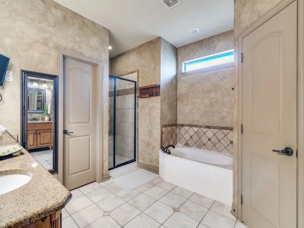 Built-in soaking tub, walk-in shower with glass enclosure, single vanity with integrated sink, patterned tile flooring, and textured wall finish
