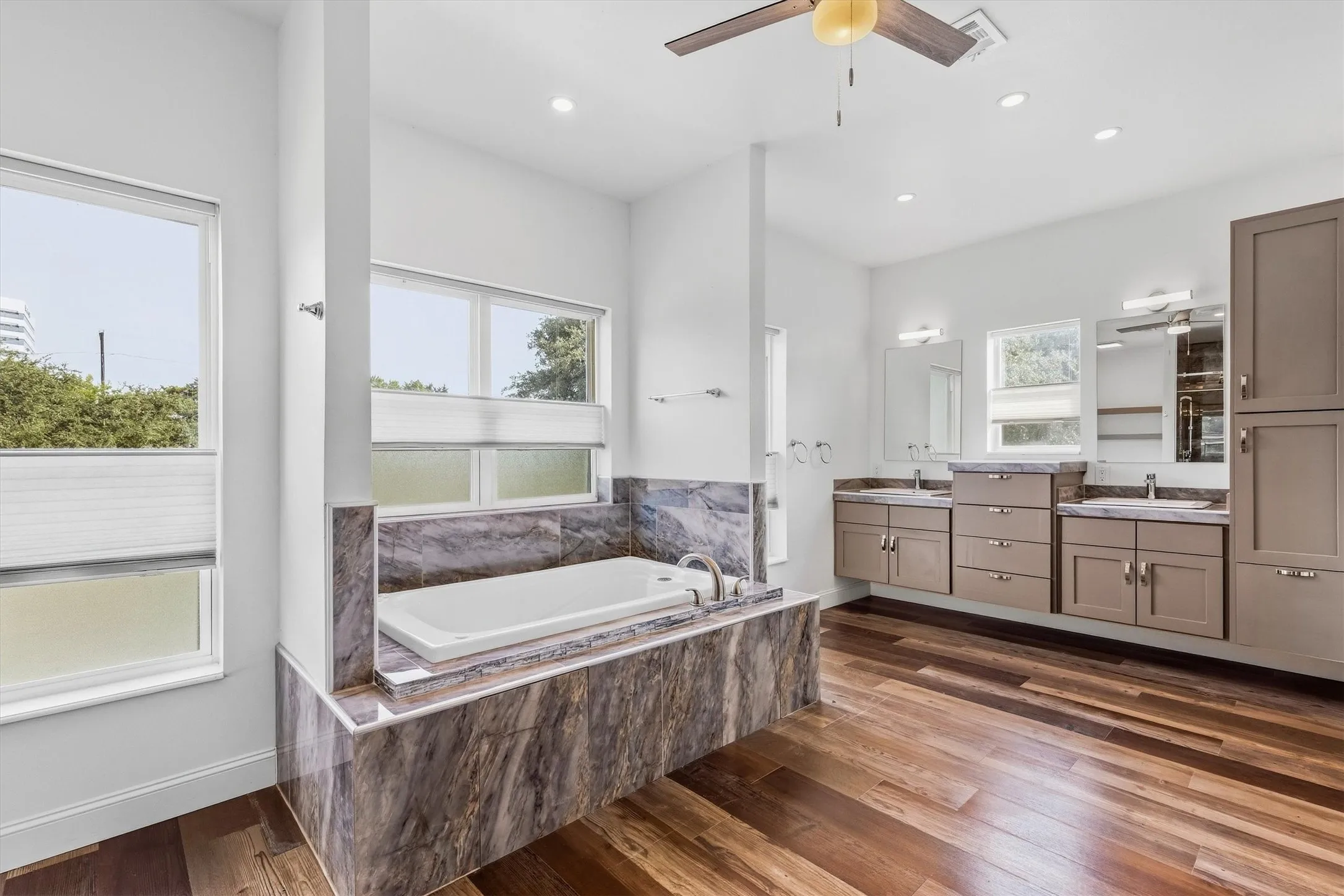 Spa-like bathroom with wood-finish flooring, a built-in tub with stone surround, dual vanities with integrated sinks, and abundant natural light from multiple windows