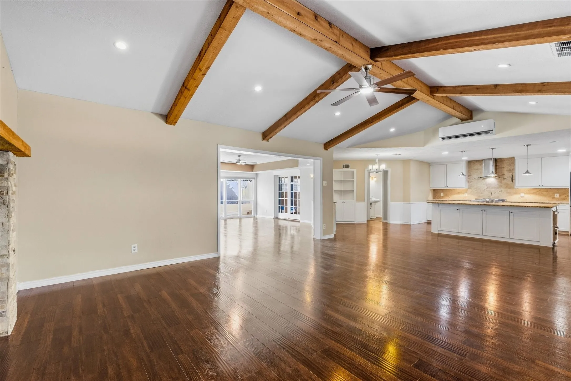 Vaulted ceiling with exposed wood beams, recessed lighting, wood-finish flooring, stone fireplace, and a kitchen featuring white cabinetry and an island with pendant lighting