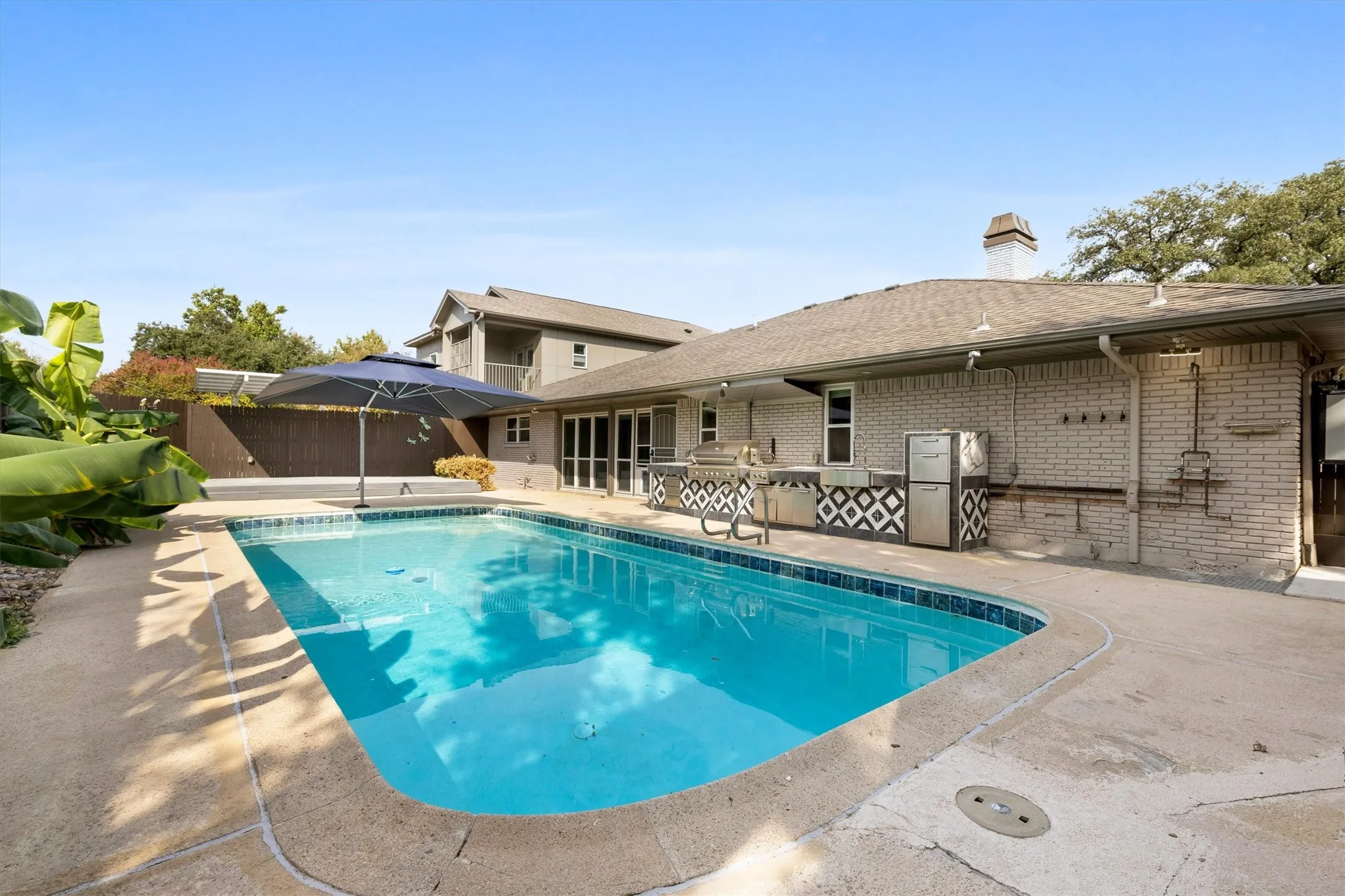 Outdoor pool with concrete surround, integrated outdoor kitchen with patterned tile accent, and a brick-clad exterior