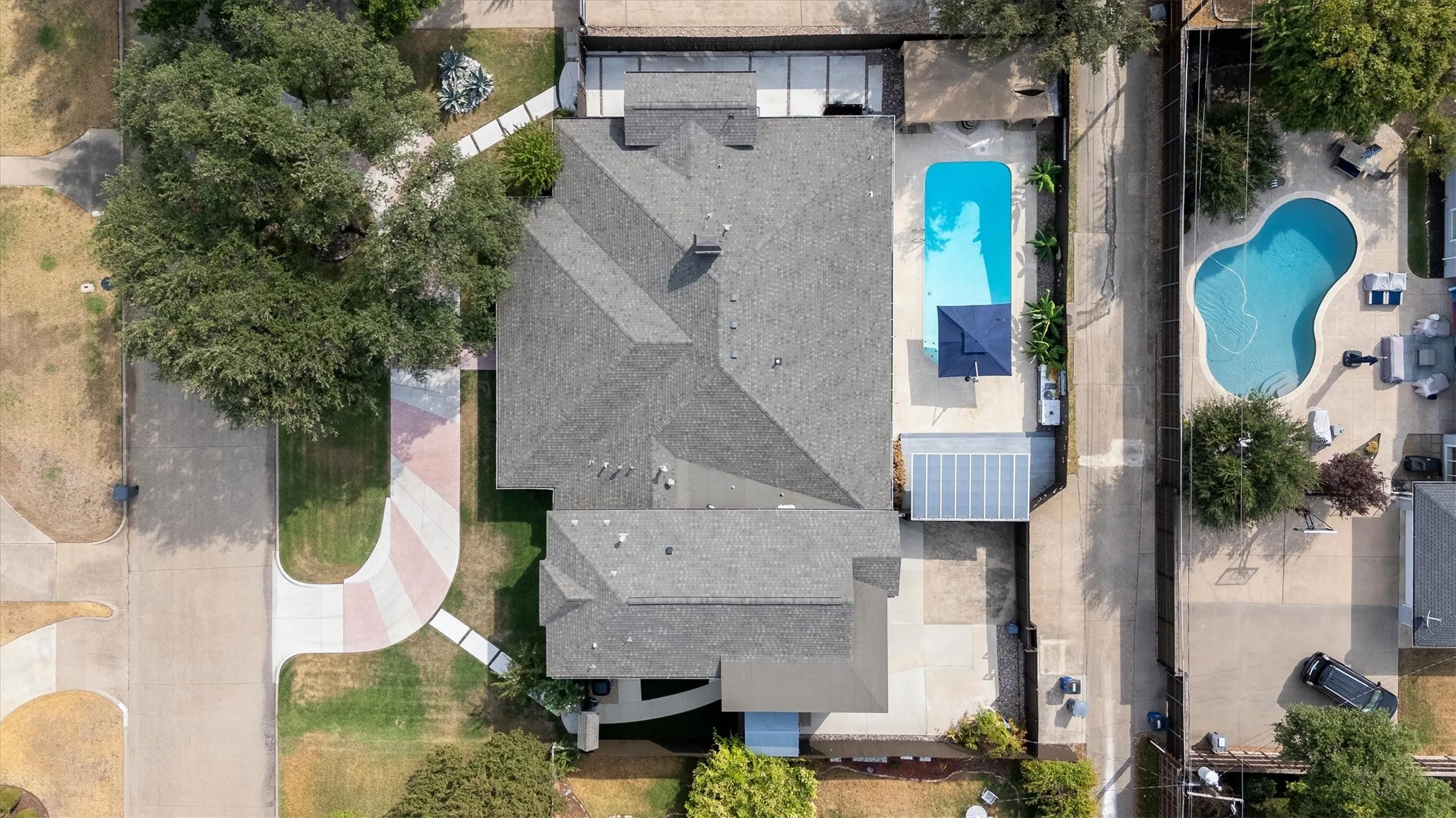Paved driveway with a patterned design, mature trees, concrete walkways, and a rectangular swimming pool with a surrounding patio