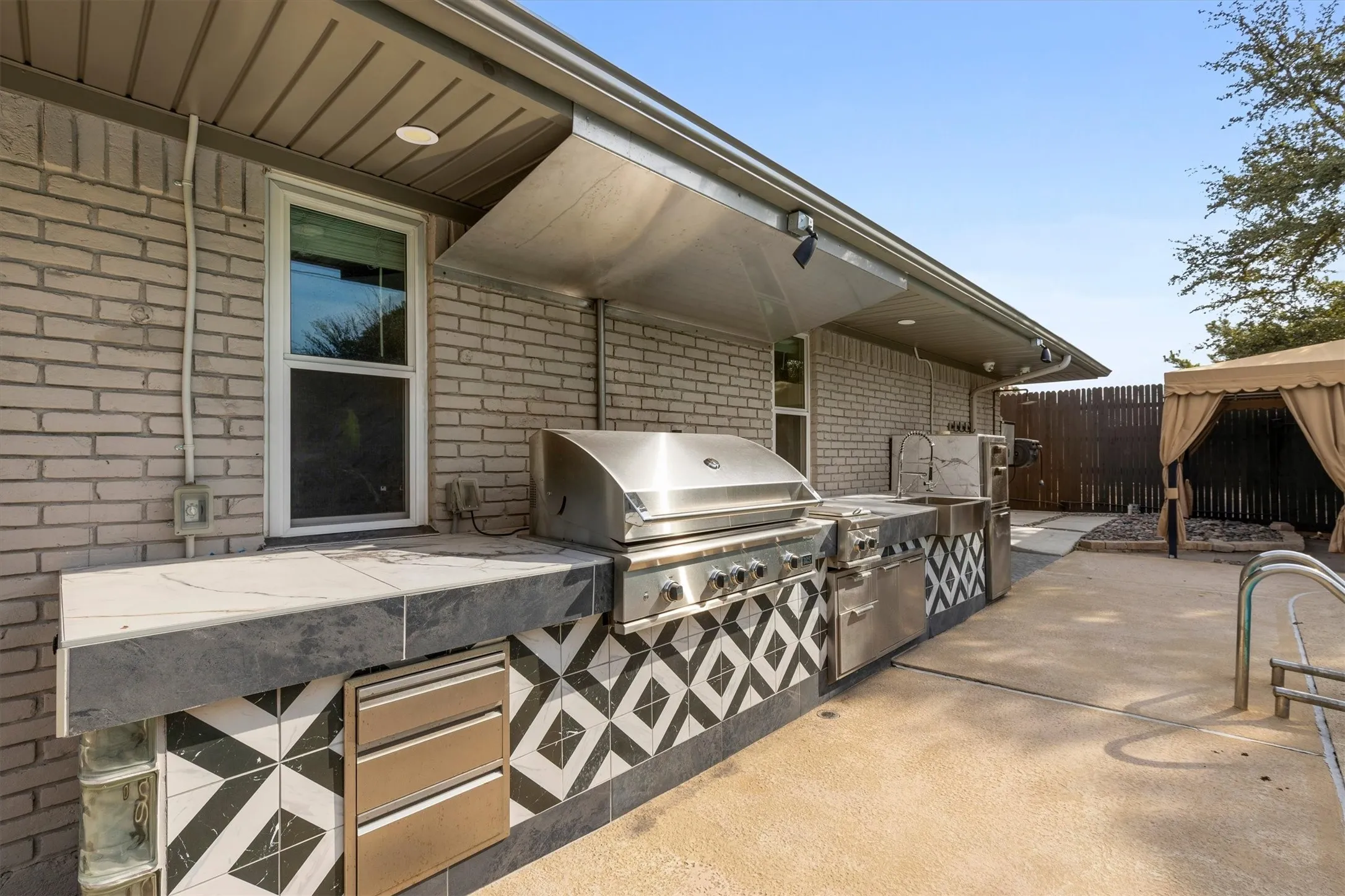 Outdoor cooking area featuring an integrated grill, stainless steel cabinetry, a built-in sink, and a patterned tile base