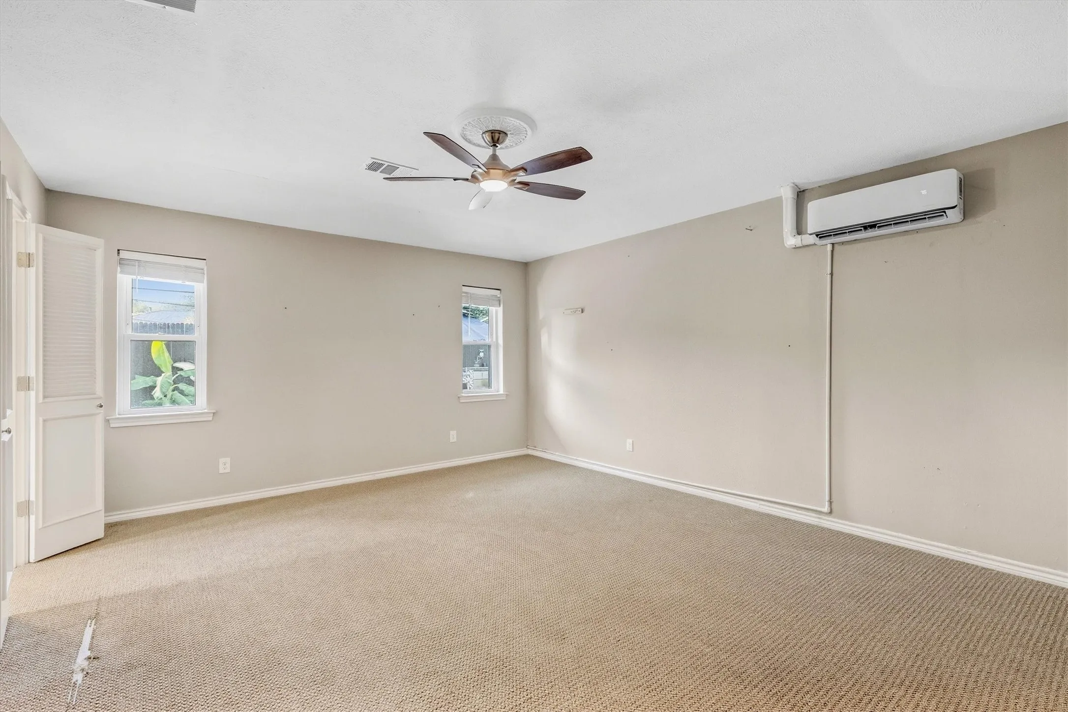 Neutral wall finishes, textured carpeting, two windows, ceiling fan with light fixture, and wall-mounted air conditioning unit