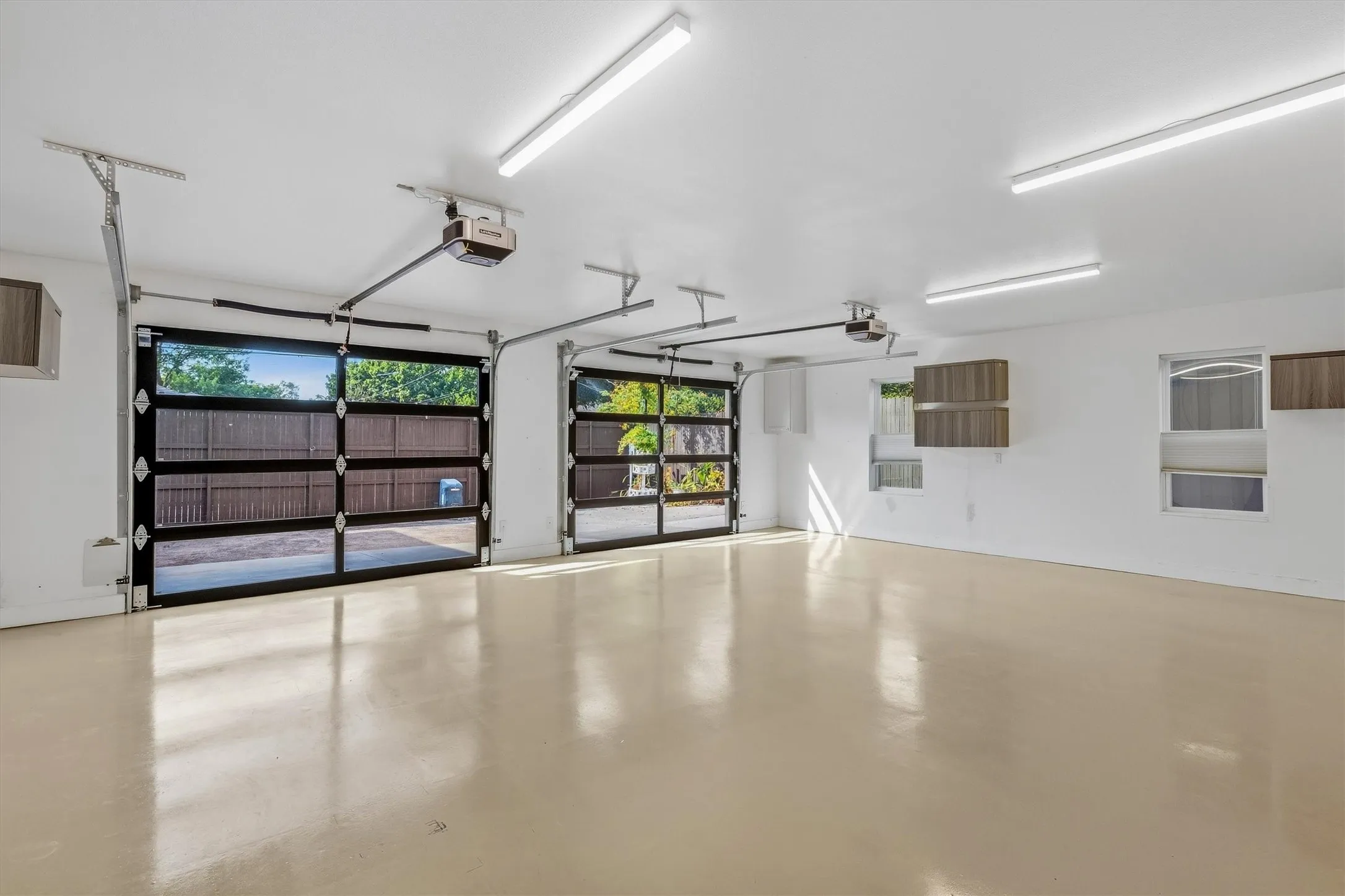 Two glass-paneled garage doors, two automatic garage door openers, multiple ceiling-mounted fluorescent light fixtures, two windows, and wall-mounted cabinetry