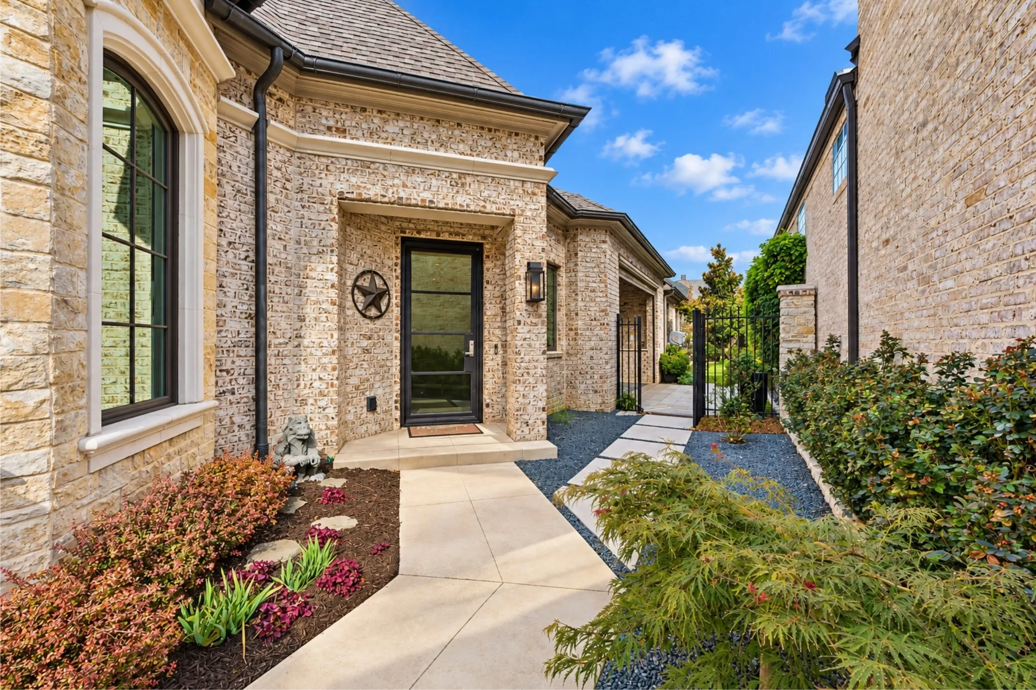 Elegant brick courtyard entry with professional hardscape paving, mature landscaping including ornamental shrubs and evergreens, arched architectural detailing, and dark-framed entry door with sidelights