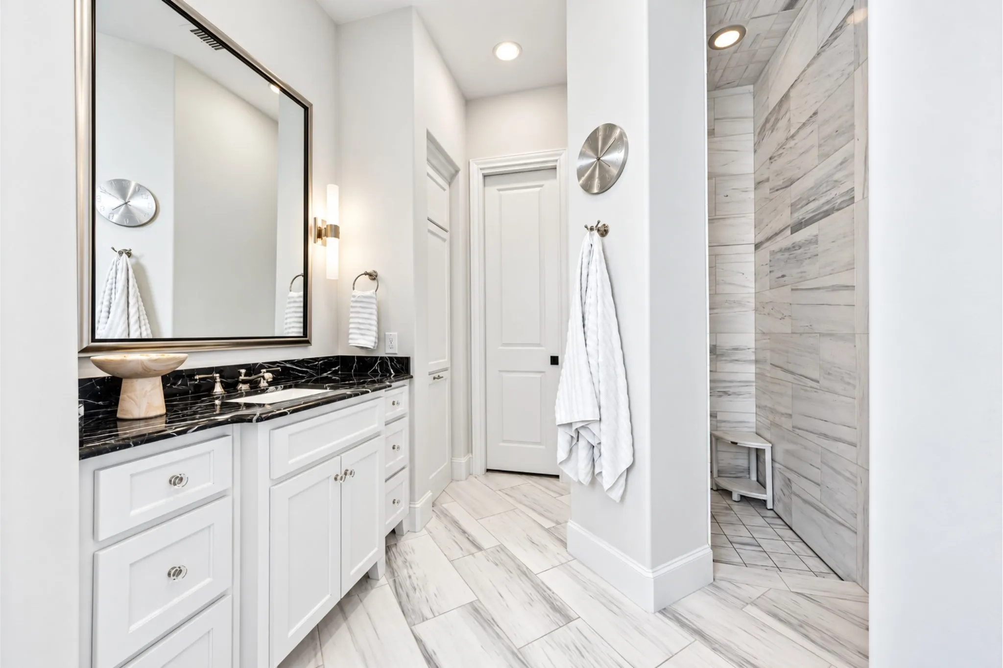 White vanity with a dark countertop and integrated sink, framed mirror with sconce lighting, and a tiled shower with a built-in bench