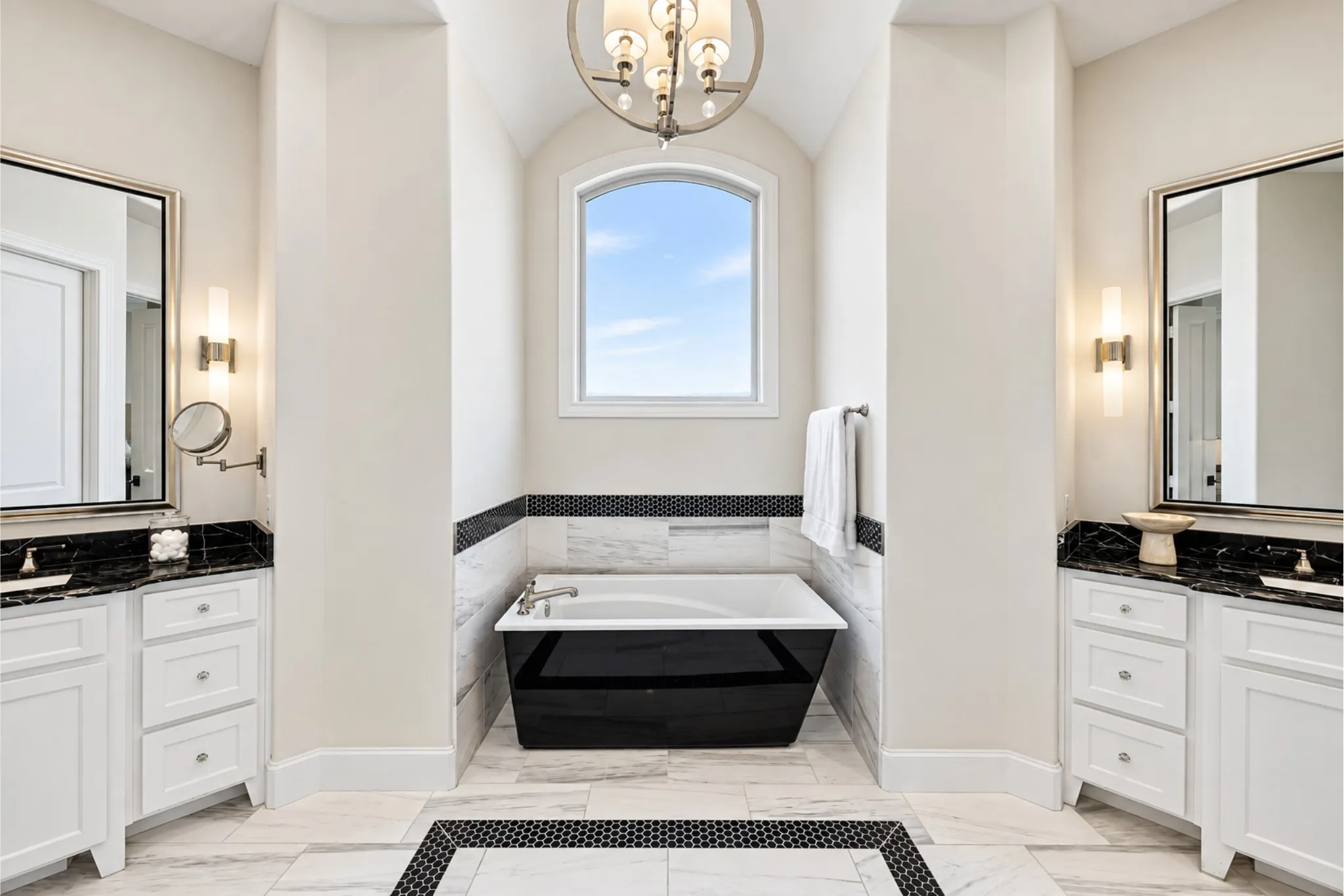 Dual vanity sinks with dark countertops, a standalone soaking tub with dark exterior, arched window, and large format tiled flooring with a mosaic inlay