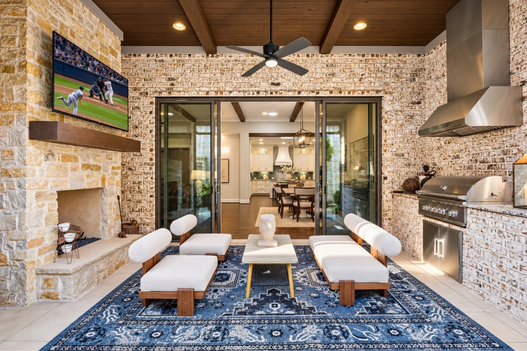 Expansive outdoor living area featuring a stone fireplace, built-in outdoor kitchen with stainless steel appliances, wood-beamed ceiling, and a ceiling fan