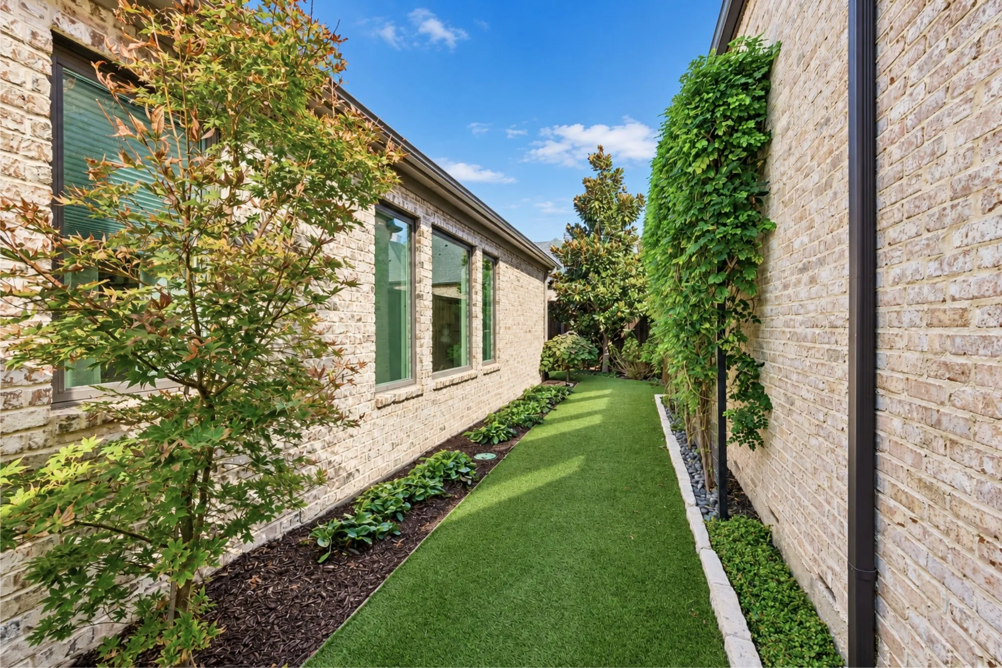 Manicured side yard featuring artificial turf, mature tree plantings, brick exterior, and vertical wall greenery