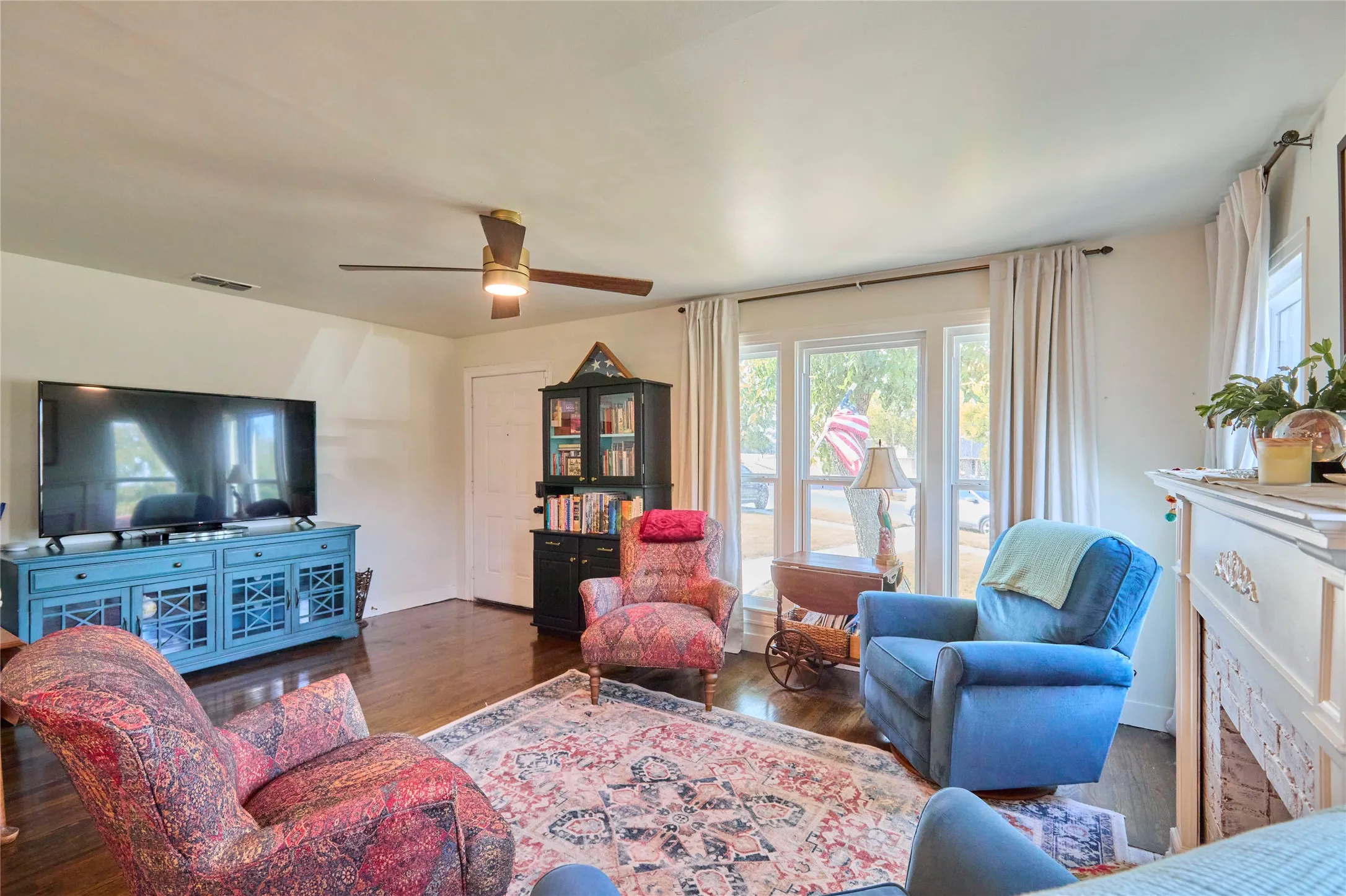 Hardwood-finish flooring, ceiling fan, multiple windows with curtains, and a decorative mantel