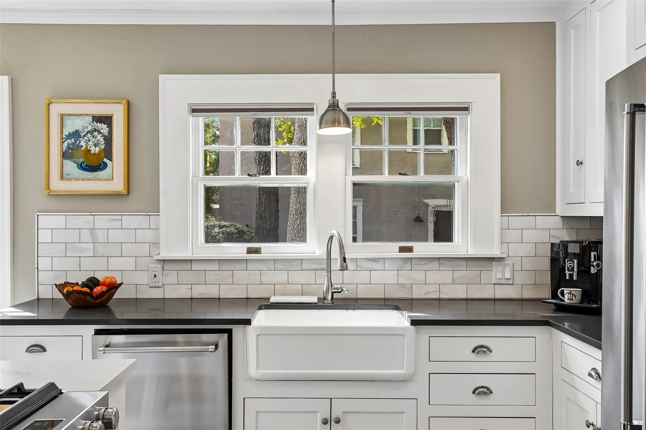 White farmhouse sink with a gooseneck faucet, dark countertops, white subway tile backsplash, white shaker-style cabinetry, and stainless steel appliances