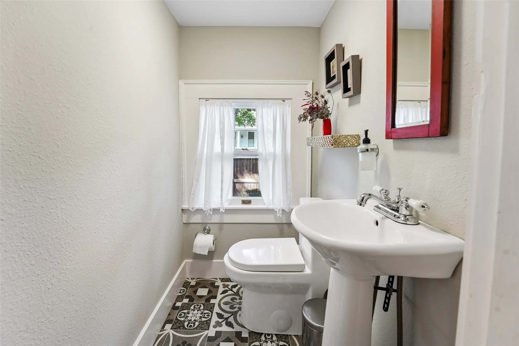 Pedestal sink with chrome faucet, toilet, window with white trim, and patterned floor tiles