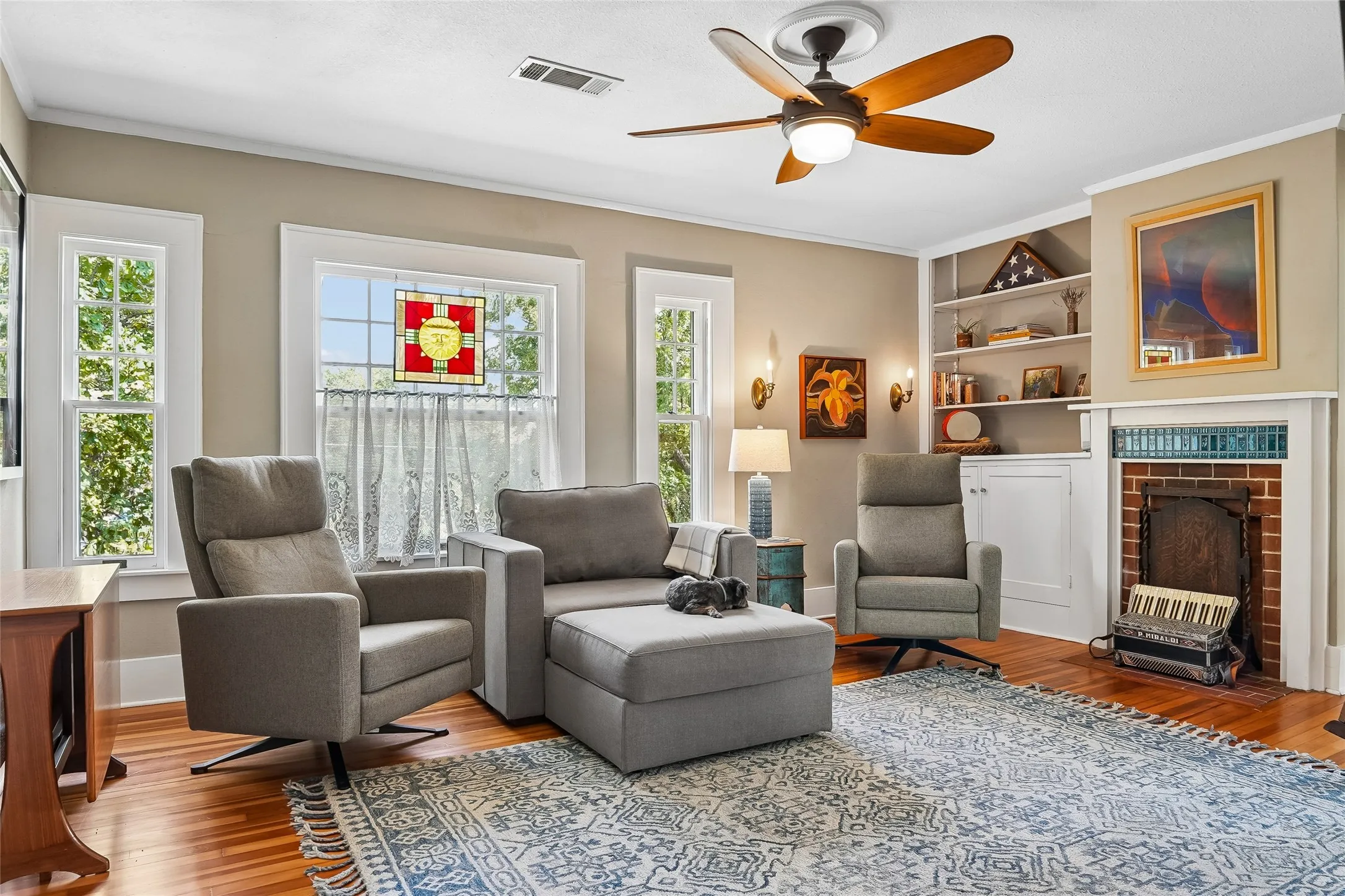 Hardwood flooring, ceiling fan with integrated lighting, built-in shelving, decorative fireplace with tiled surround, and multiple windows with trim