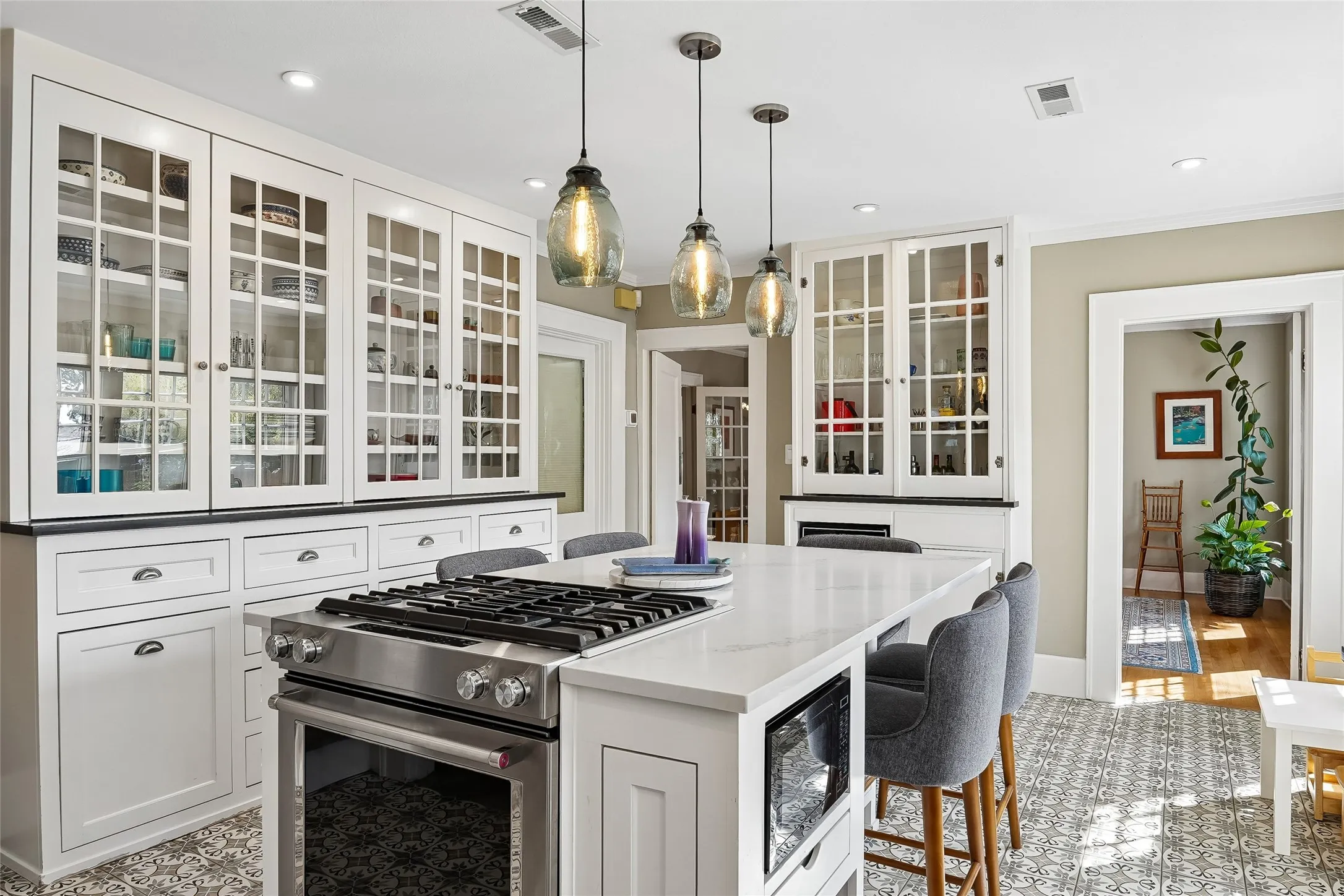Gourmet kitchen featuring a center island with built-in stainless steel range, white shaker cabinetry with glass-front uppers, and patterned tile flooring