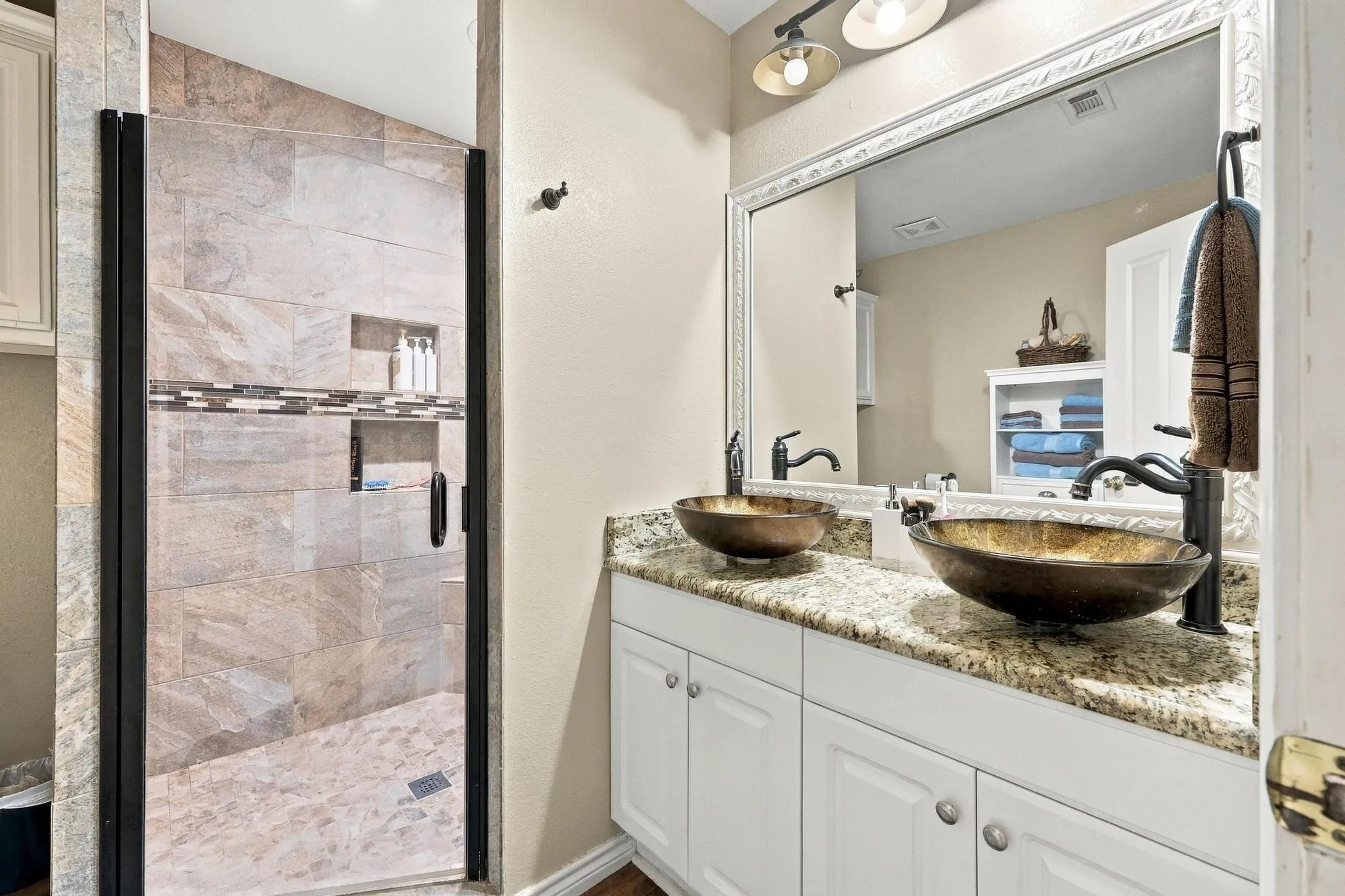 Dual vessel sinks with bronze-finish fixtures, granite countertop vanity, decorative framed mirror, and a walk-in shower with tile surround and built-in niches