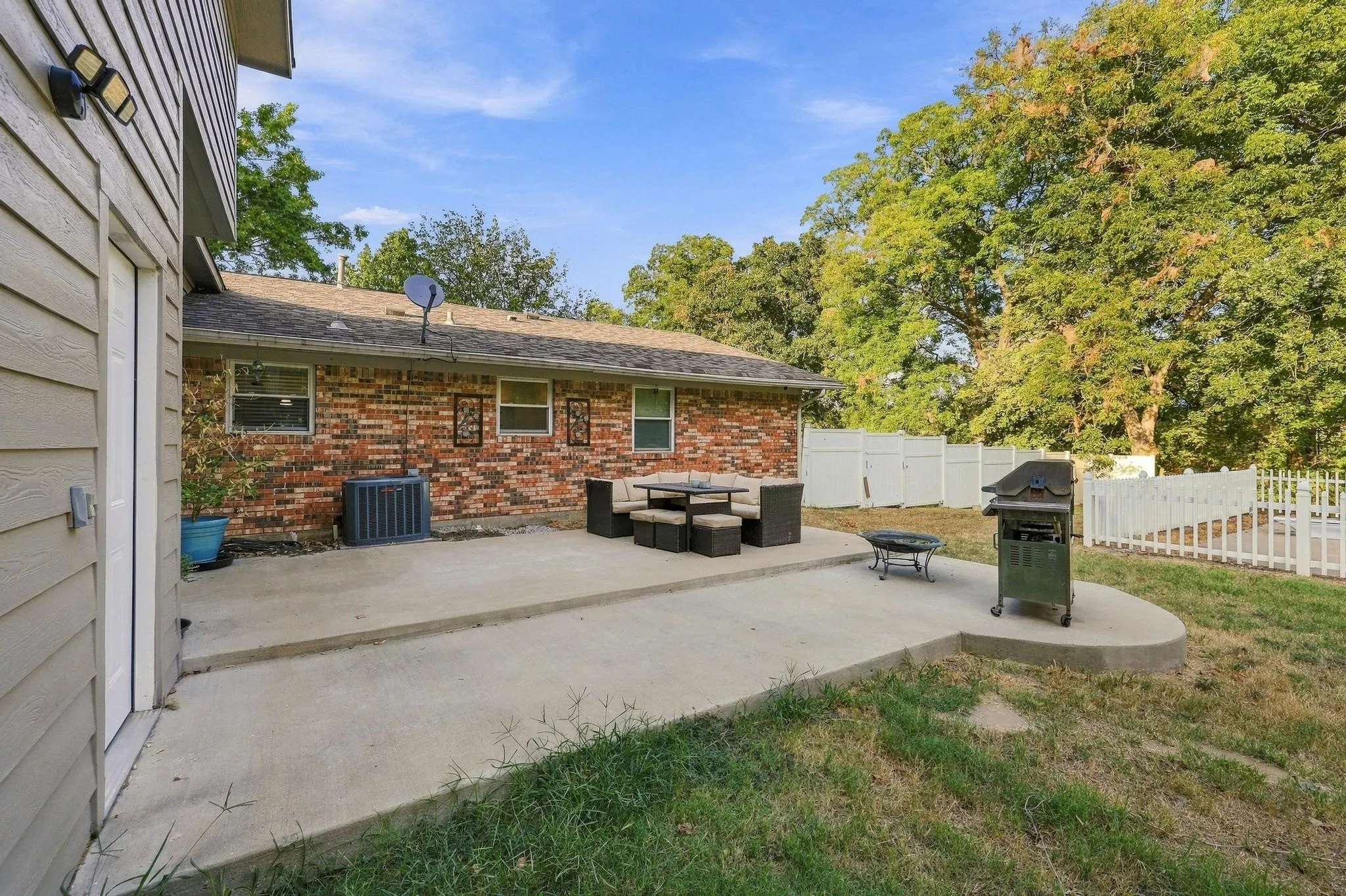 Expansive concrete patio with a brick exterior, surrounded by mature trees and a white vinyl fence