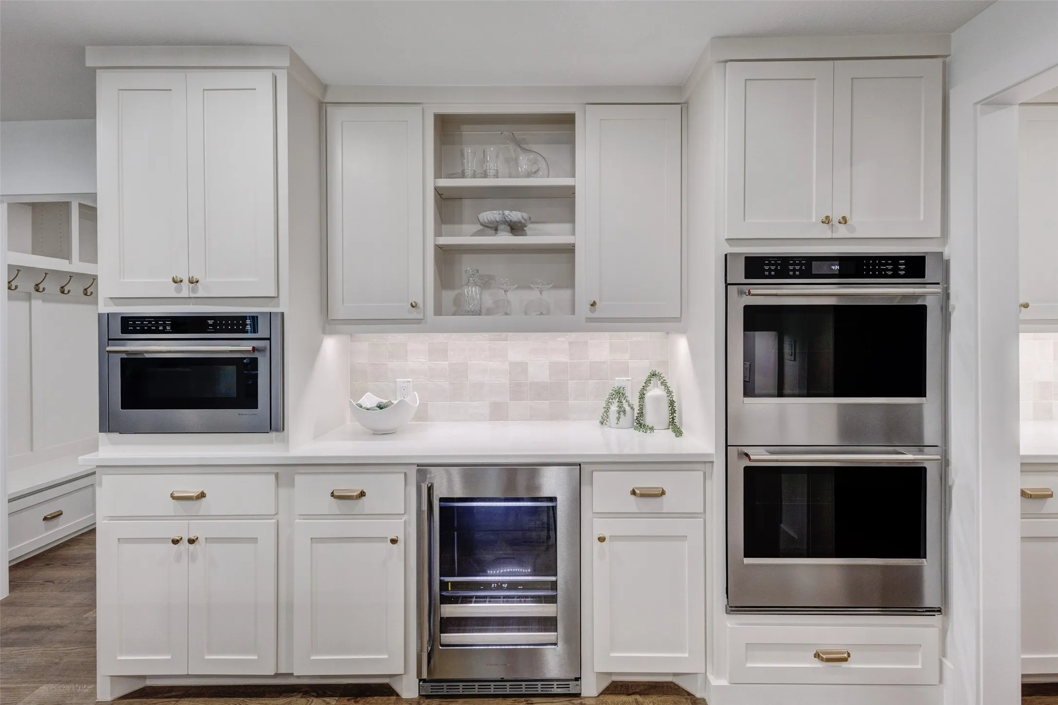 White shaker-style cabinetry with gold-tone hardware, stainless steel built-in microwave, integrated beverage cooler, and double wall ovens
