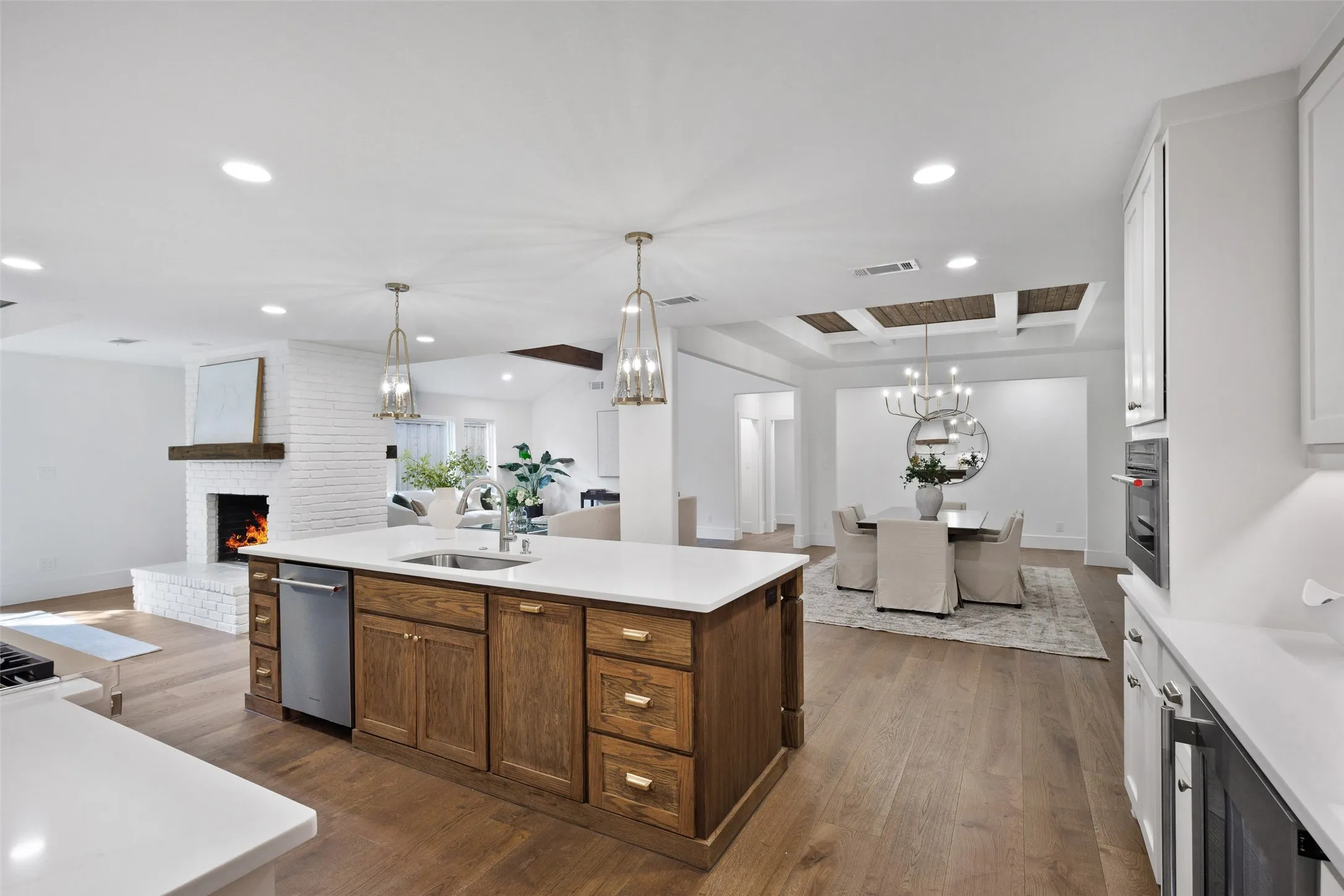 White brick fireplace with wood mantel, hardwood flooring, coffered ceiling, white cabinetry, and a kitchen island with storage