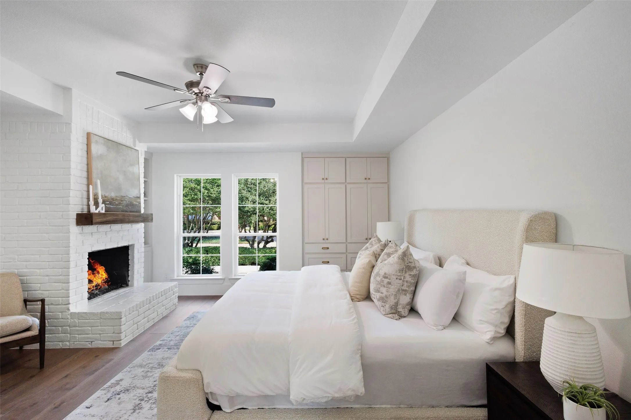 Recessed ceiling with an overhead fan, white painted brick fireplace with a wood mantle, two large windows, wood-finish flooring, and built-in cabinetry