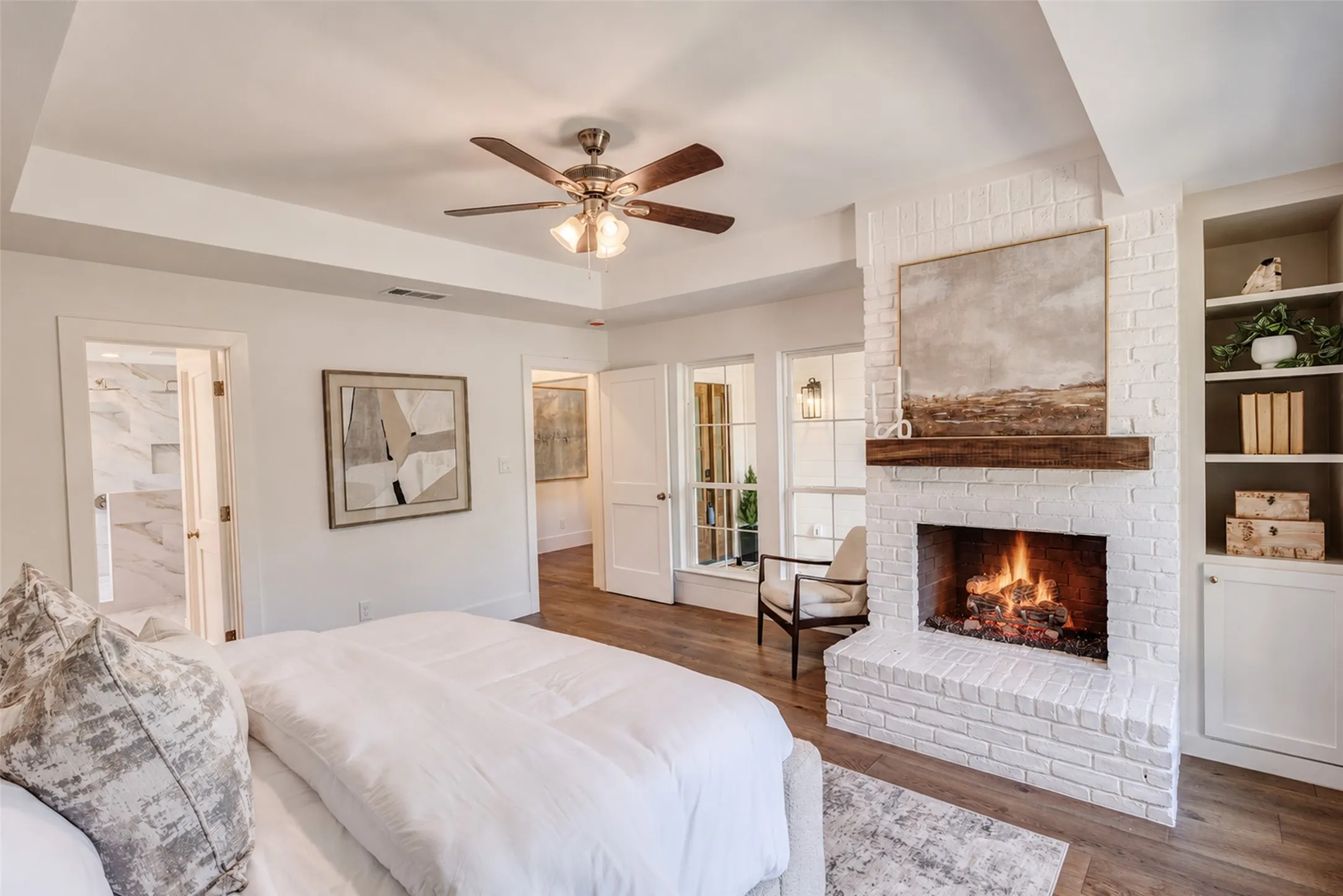 Tray ceiling with a ceiling fan, wood-finish flooring, white brick fireplace with a wood mantle, built-in shelving, and an adjacent bathroom with stone-finish walls