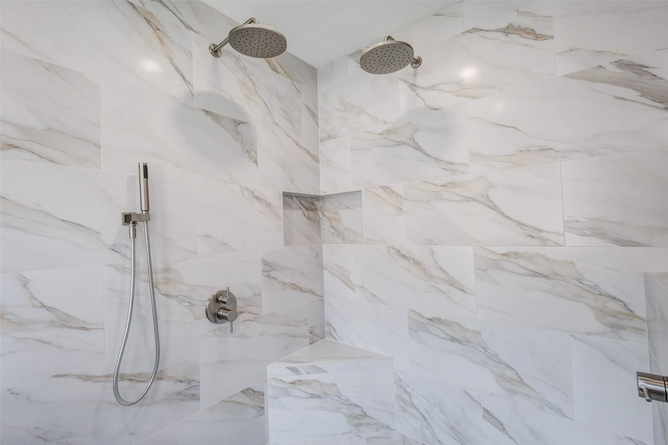 White and gold-veined wall tile, dual rainfall showerheads, handheld shower sprayer, built-in corner shelf, and brushed nickel fixtures