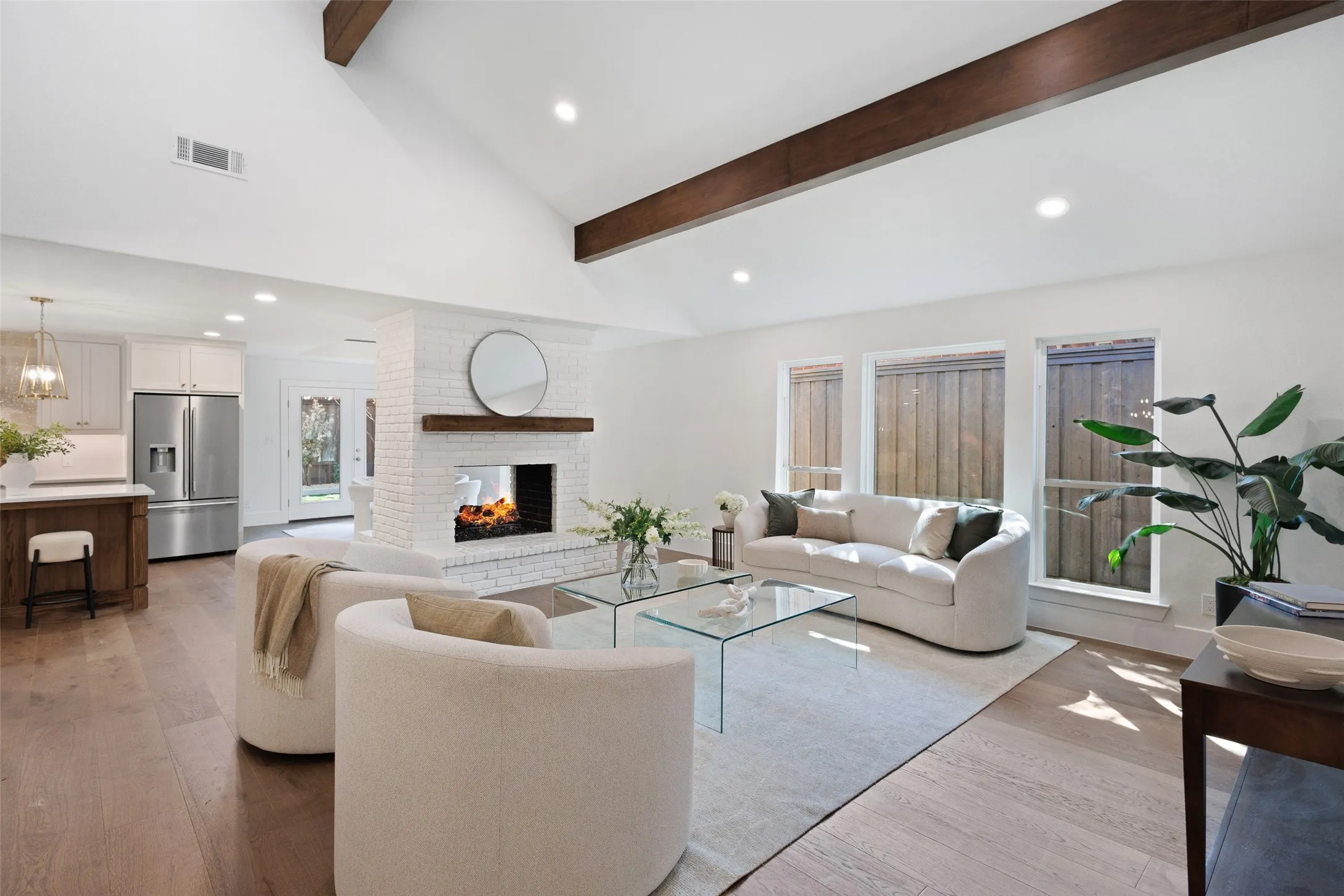 Vaulted ceiling with exposed wood beams, wood-finish flooring, white brick fireplace with wood mantel, recessed lighting, and stainless steel refrigerator