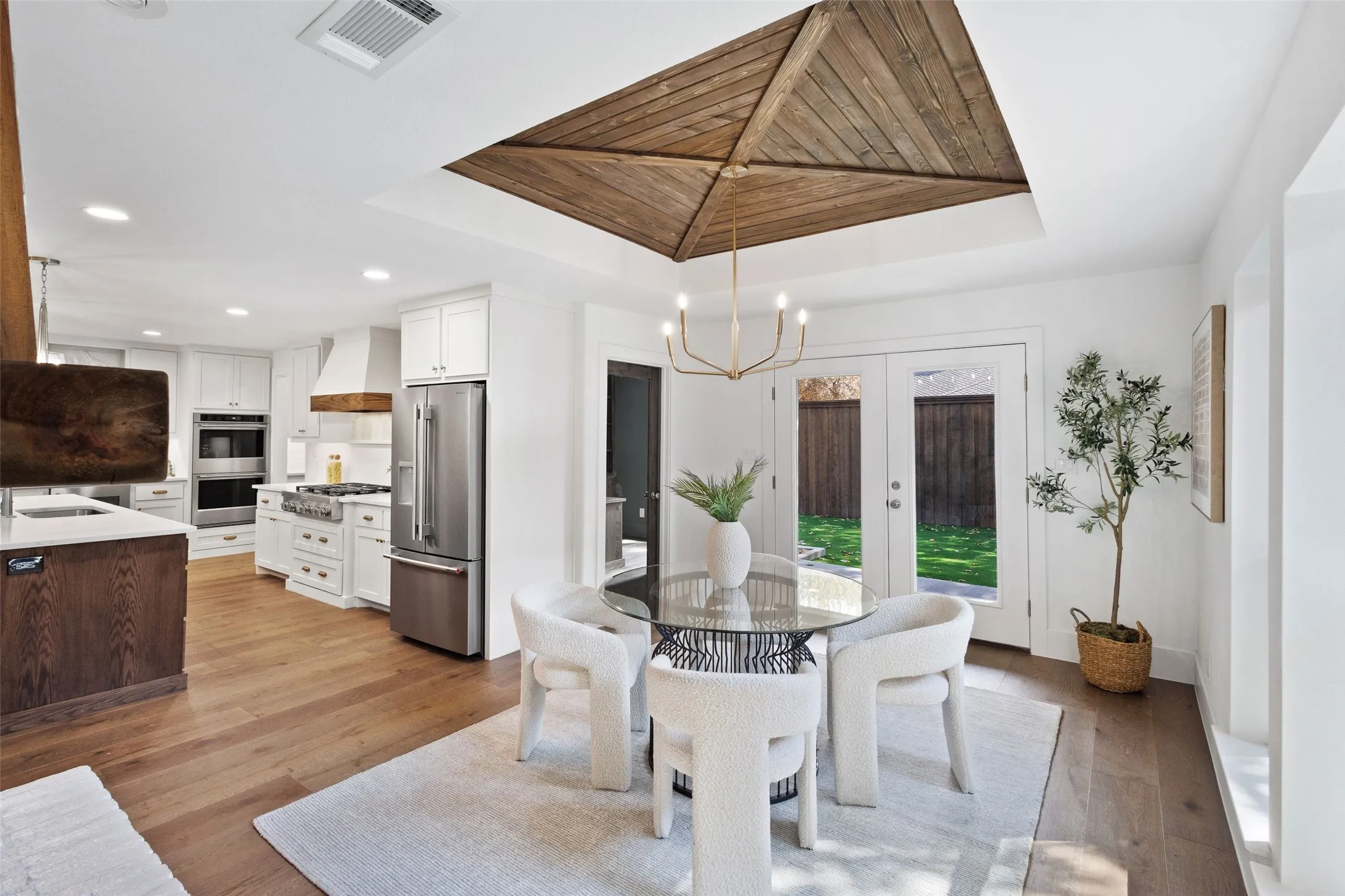 Wood-finish flooring throughout, kitchen with white cabinetry and stainless steel appliances, dining area featuring a wood-panel ceiling design, and double doors opening to an outdoor area